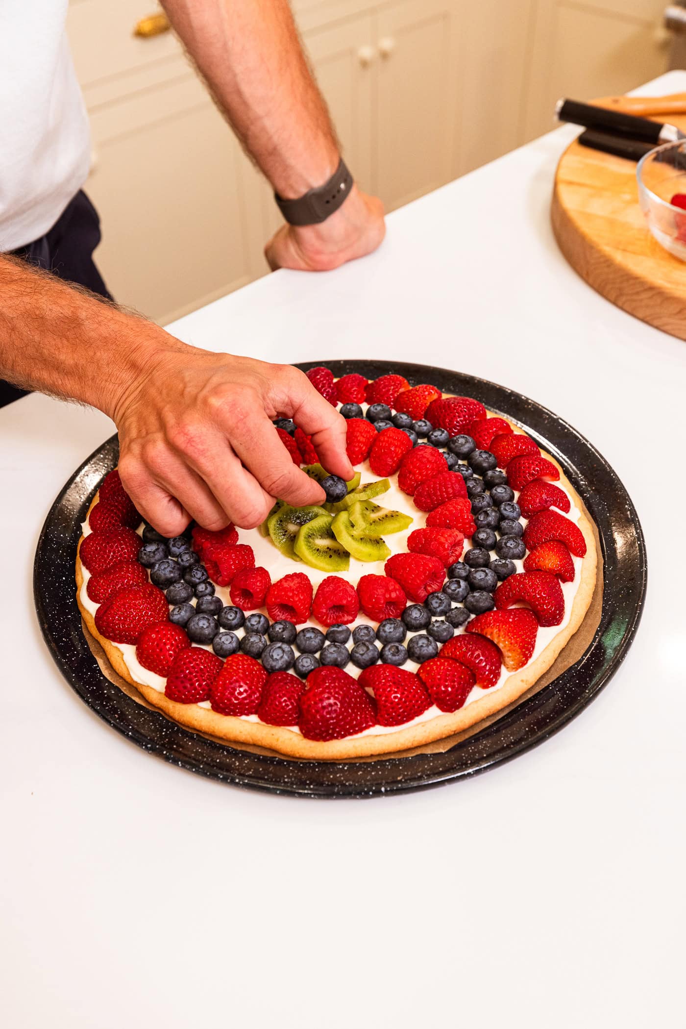Hand placing fruit on top of a fruit pizza on a baking dish.