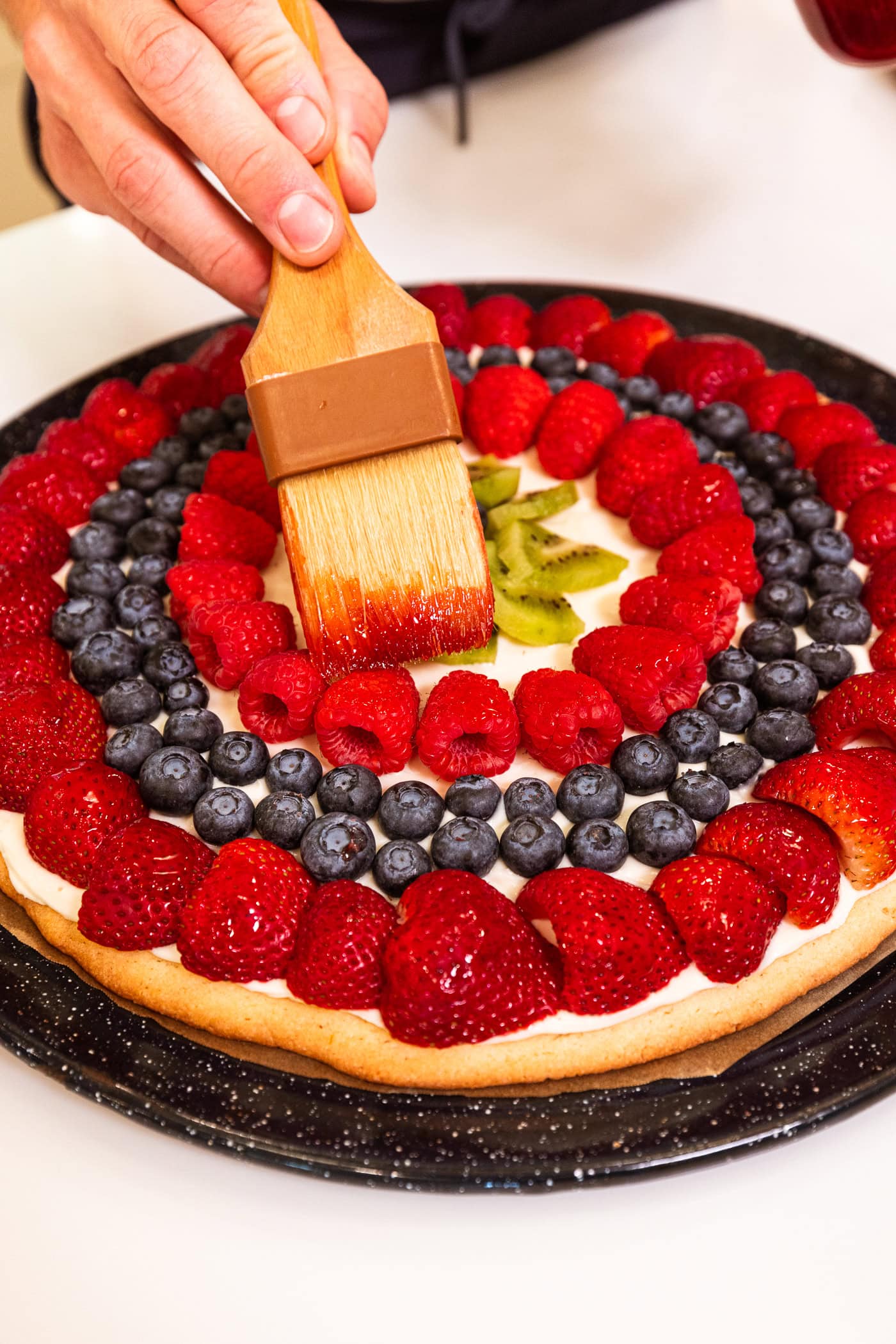 Hand using brush to brush on a jam glaze on top of a fruit pizza.