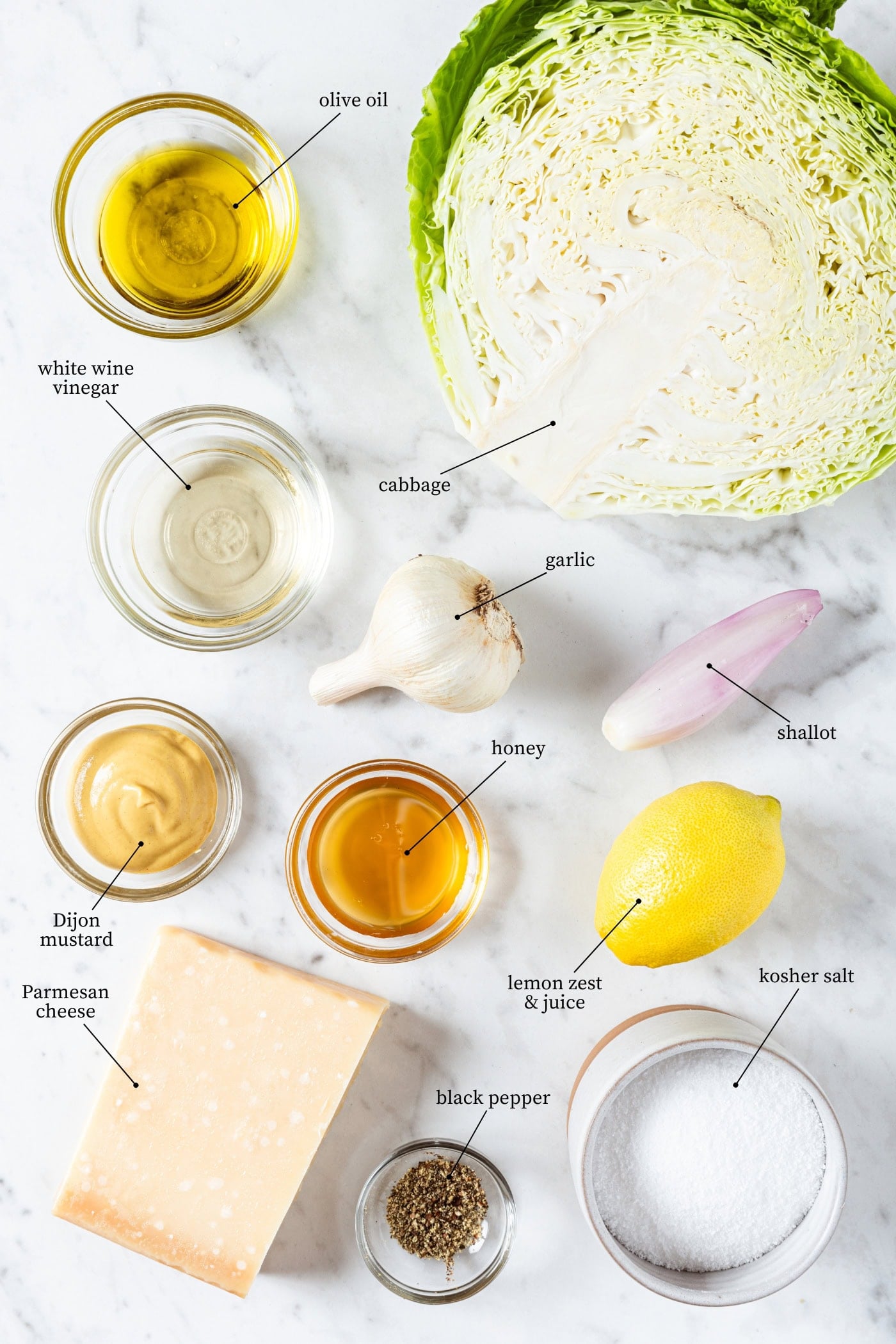 White marble surface filled with ingredients needed to make grilled cabbage salad including cabbage, oil, mustard, honey, lemon, Parmesan cheese, and more.