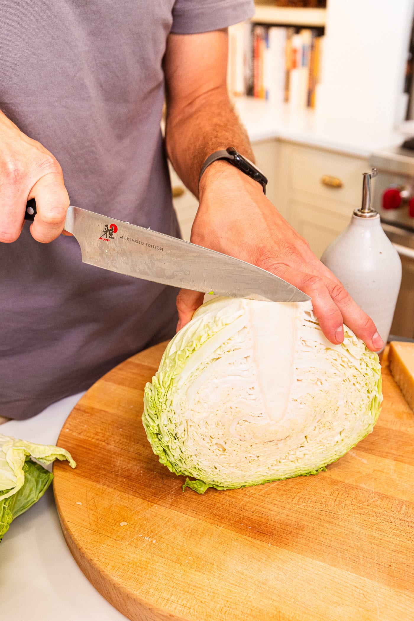 Hand holding a whole cabbage using a knife to slice it in half on a wooden cutting board.