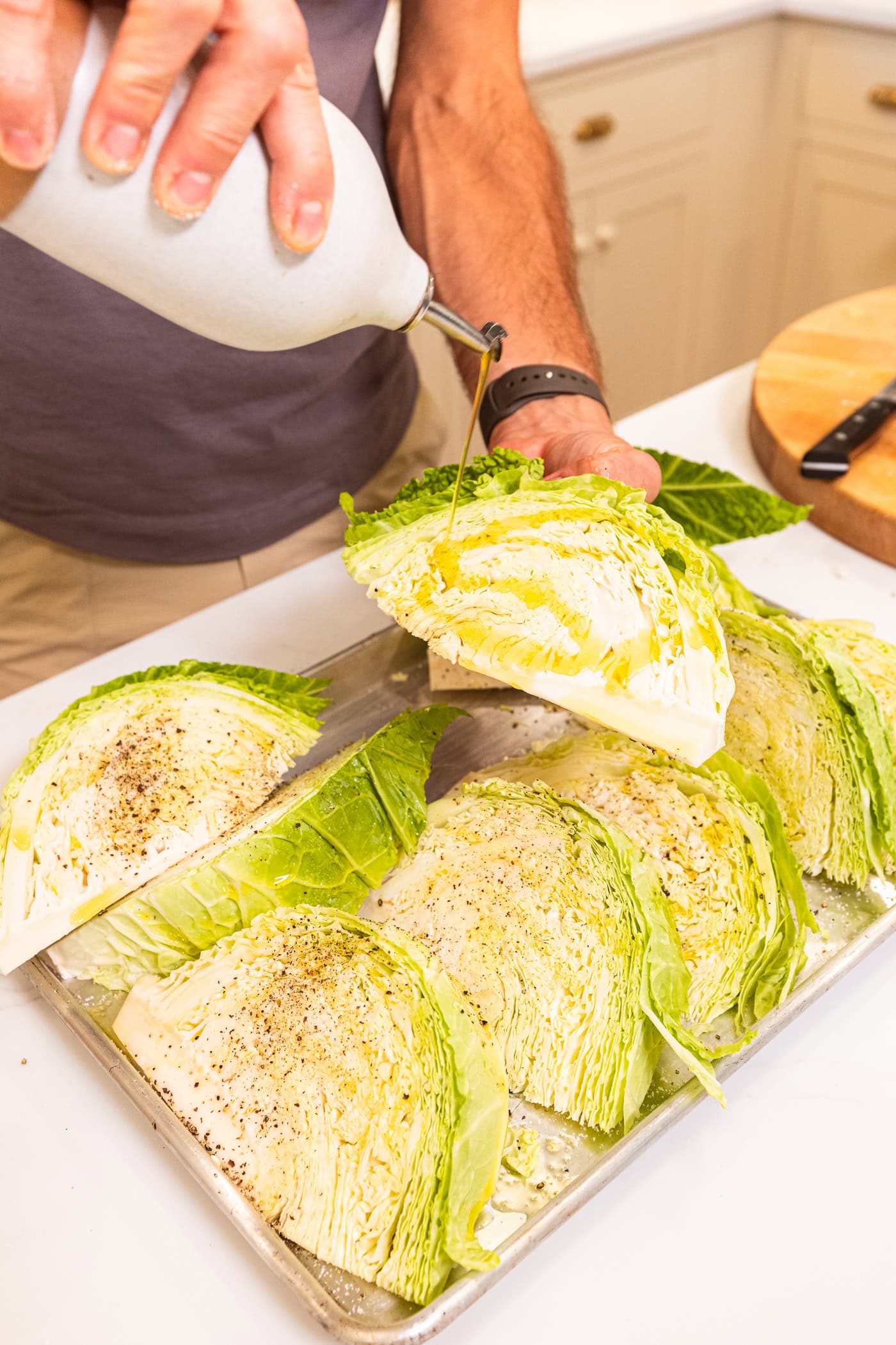 Hand using oil to drizzle over the top of cabbage sliced into pieces on a metal baking pan.