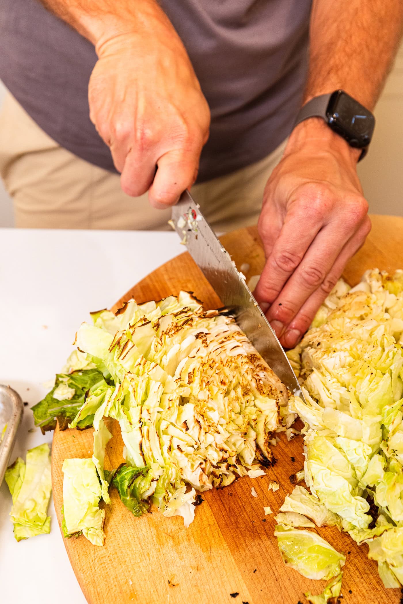 Hand using knife to chop up grilled cabbage wedges on a wood cutting board.