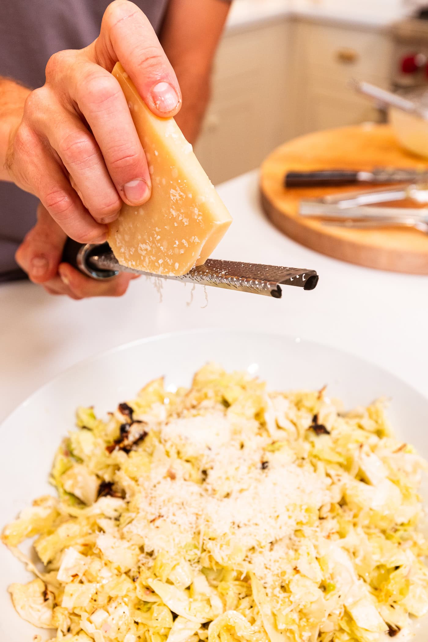 Hand using grater to grate Parmesan cheese on top of chopped and grilled cabbage in large white bowl.