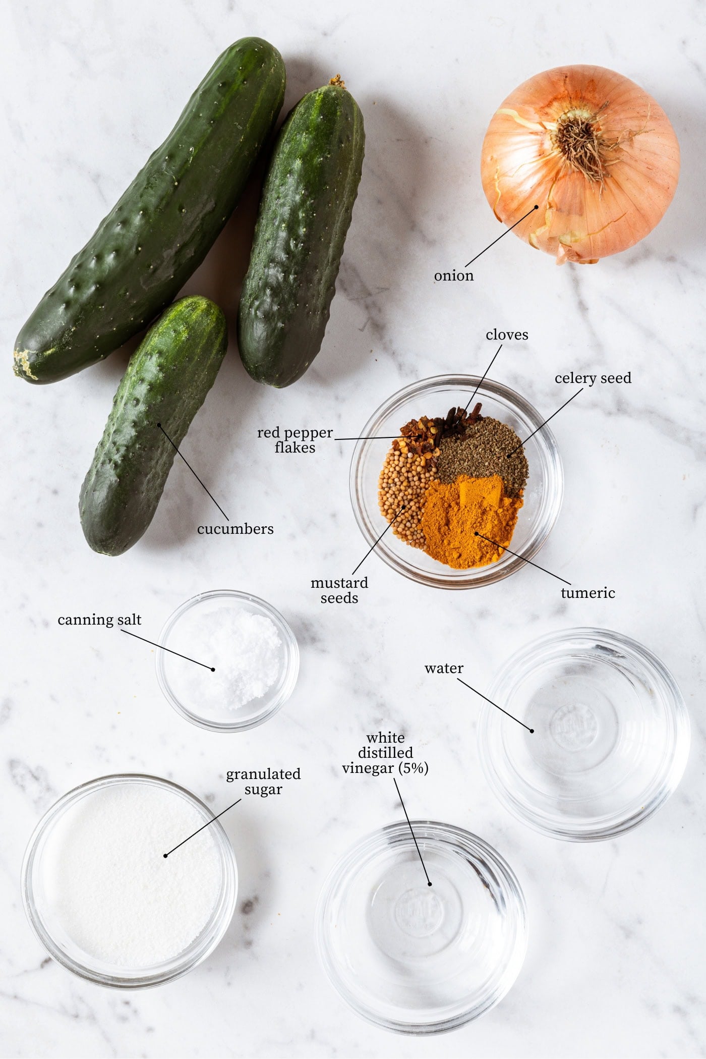 A marble surface with ingredients needed to can bread and butter pickles including sugar, vinegar, water, canning salt, cucumbers, onion, and spices.
