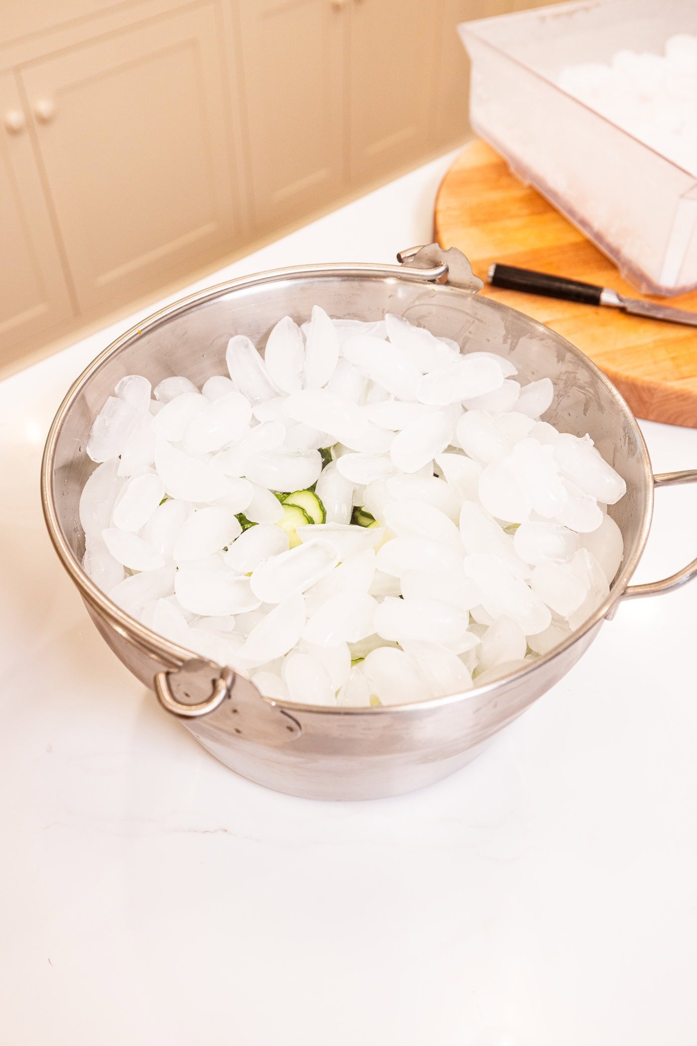 Large metal kettle with ice cubes all over the top of cucumber slices.