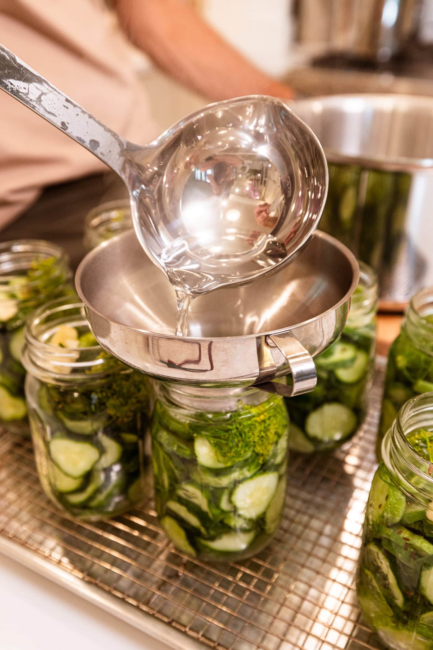 Ladle pouring vinegar brine into jars filled with sliced cucumbers for pickles.