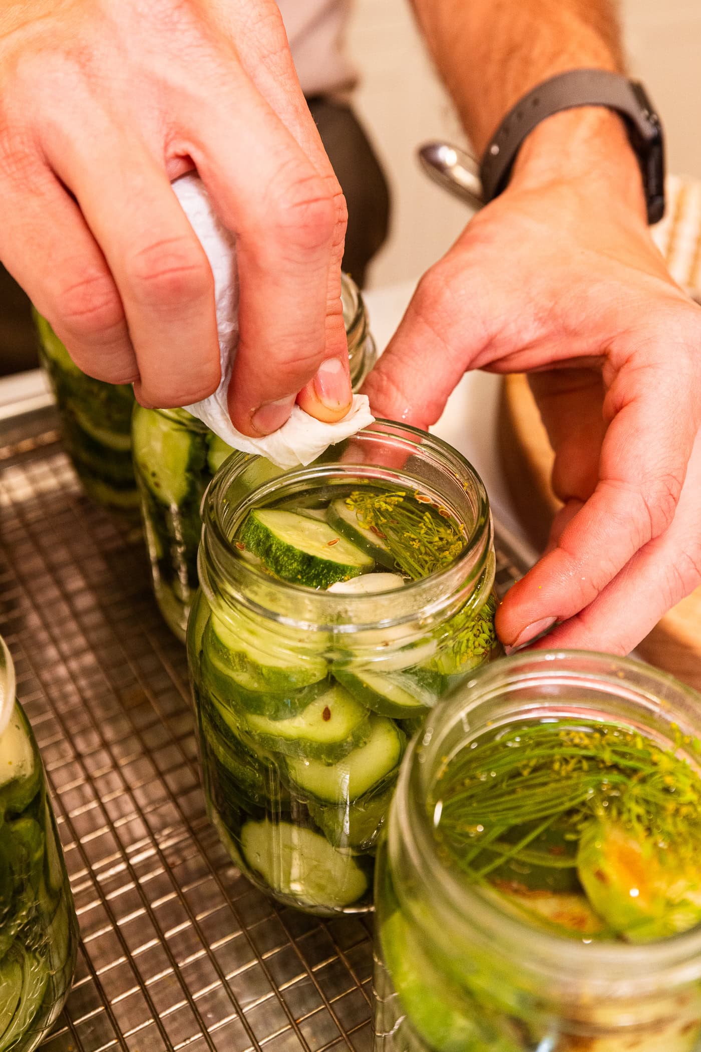 Hand using piece of paper towel to wipe off the rim of a jar filled with cucumber slices.