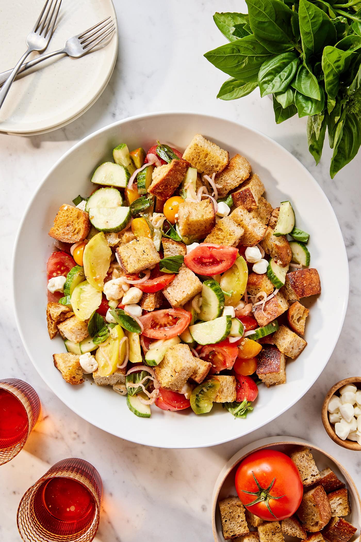 Large white bowl filled with panzanella salad all on white surface with plates and fresh basil around.