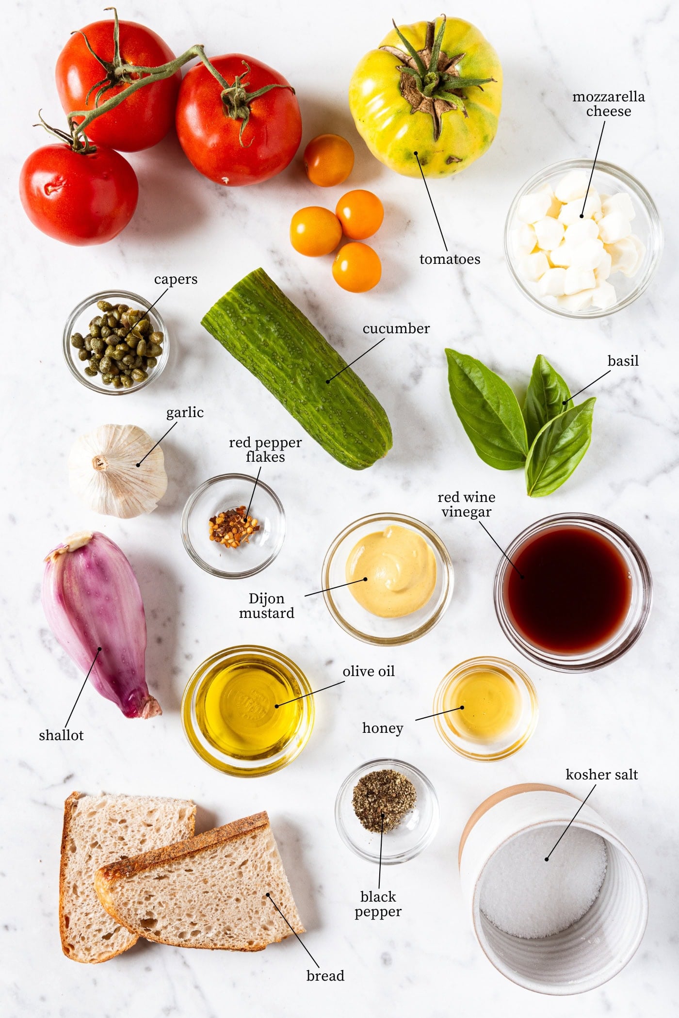 White marble surface filled with ingredients for panzanella including tomatoes, cucumber, basil, mozzarella, bread, and more.