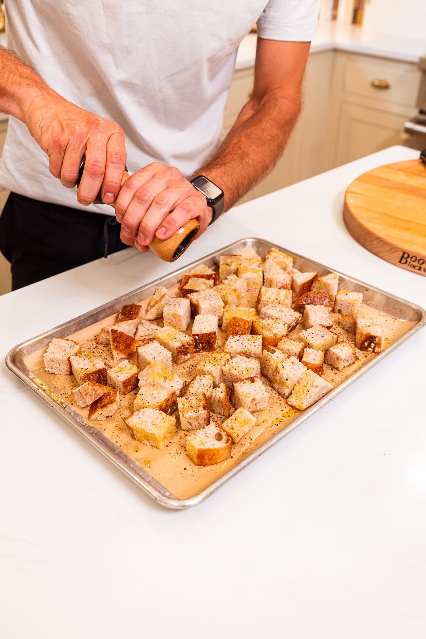 Hand using grinder to put black pepper on top of bread cubes on a sheet pan before going into the oven.