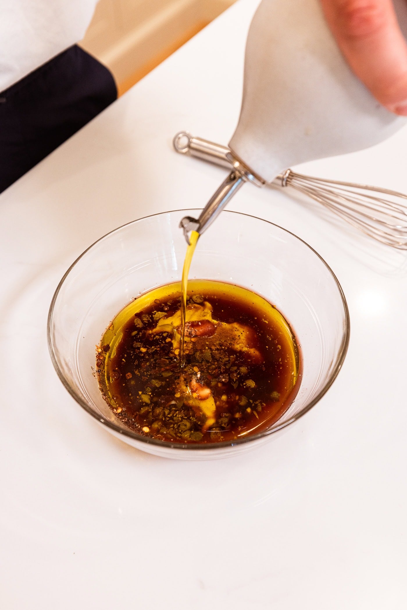 Glass bowl filled with ingredients needed for a vinaigrette with olive oil being drizzled into the bowl.