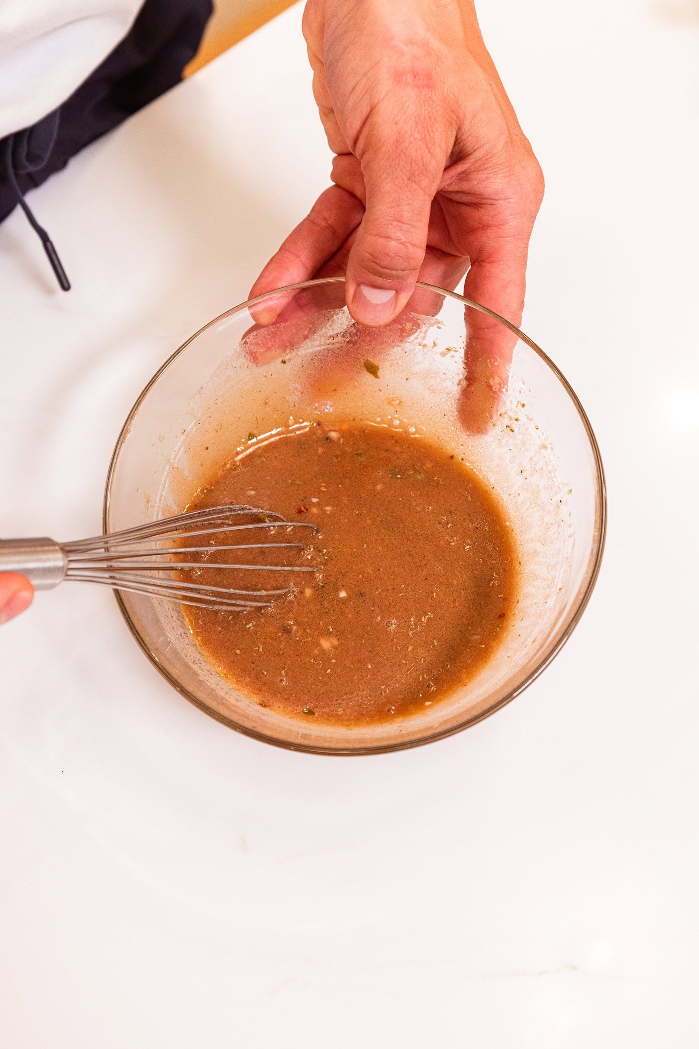 Glass bowl filled with brown-colored vinaigrette all on white surface after being whisked together.