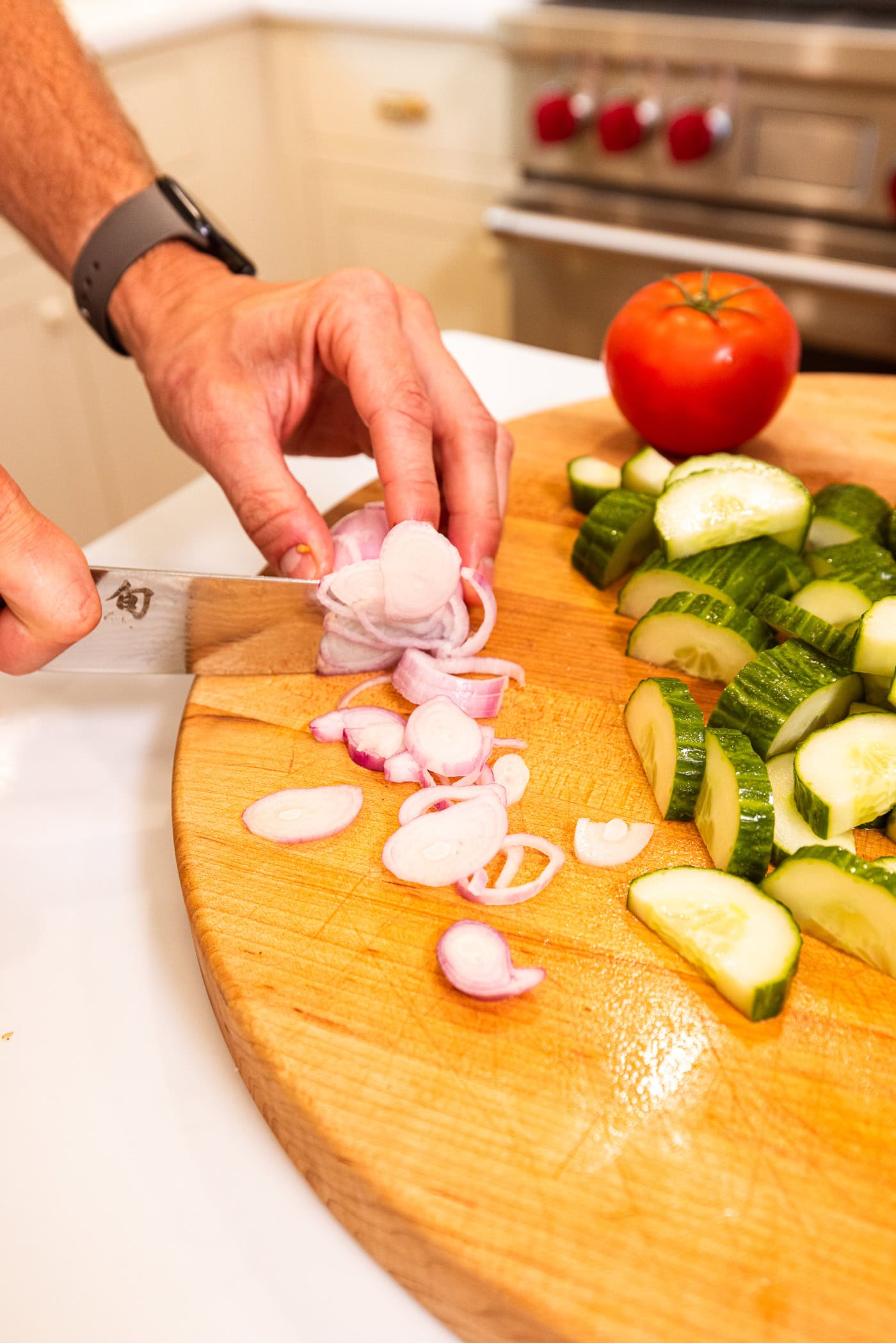 Hand slicing shallot into thin strips on wooden cutting board with sliced cucumber.