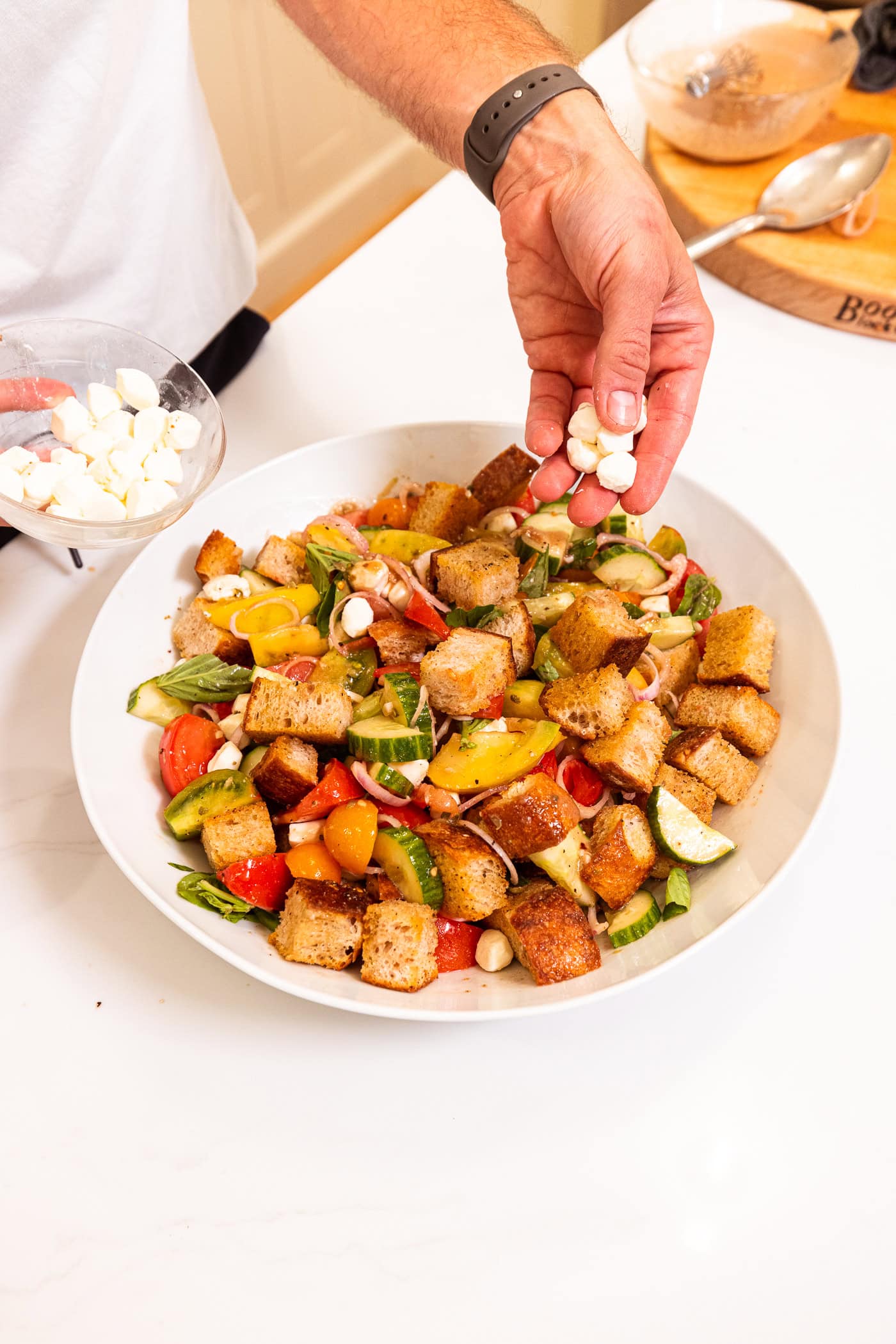 Hand placing pieces of mozzarella cheese on top of tomatoes, cucumbers, and crusty bread in a large white bowl.
