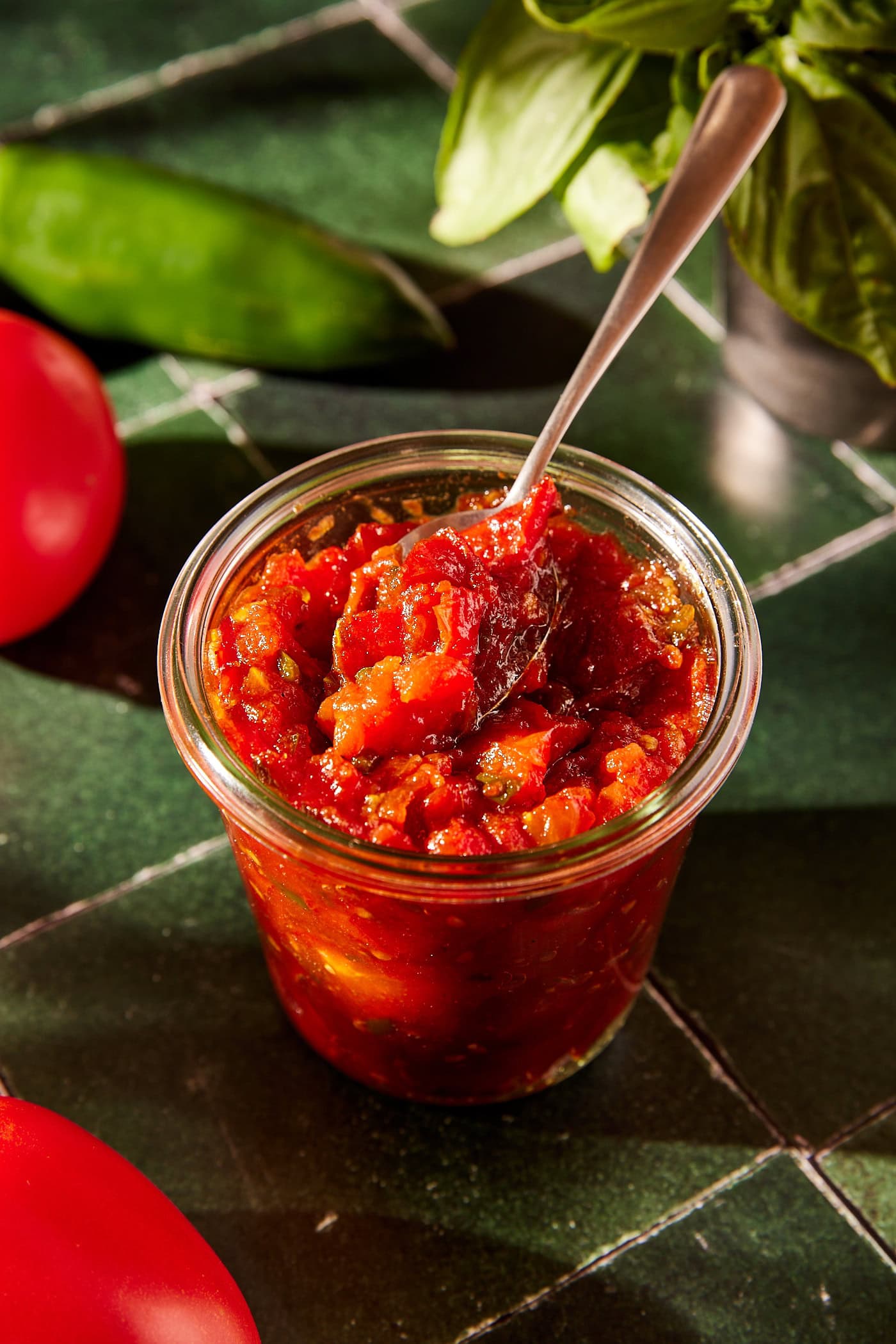 Spoon scooping out tablespoon of tomato jam from a glass jar filled with more with tomatoes and basil all around on a green tile surface.