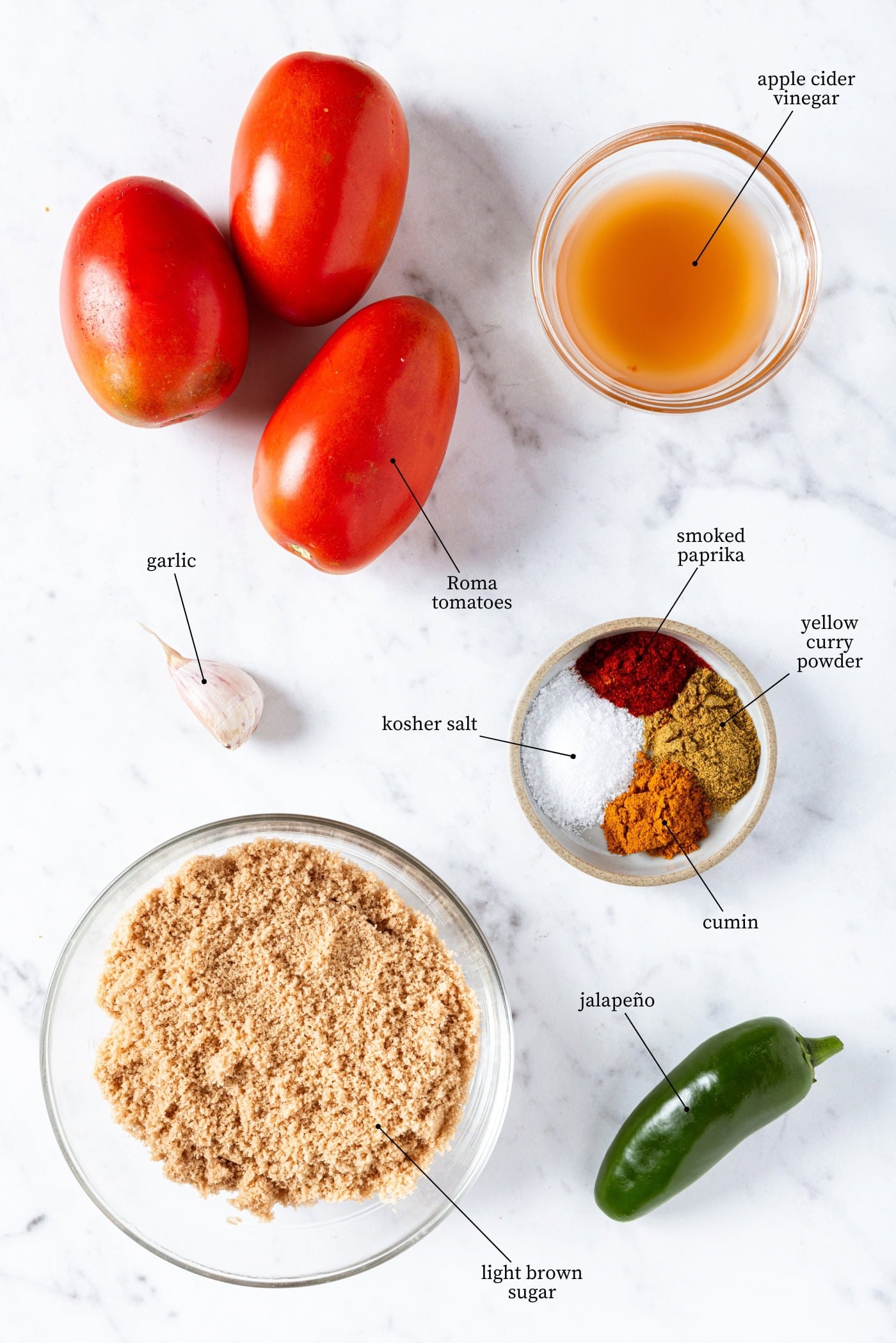 White marble surface with ingredients needed to make tomato jam recipe including tomatoes, brown sugar, spices, and more.