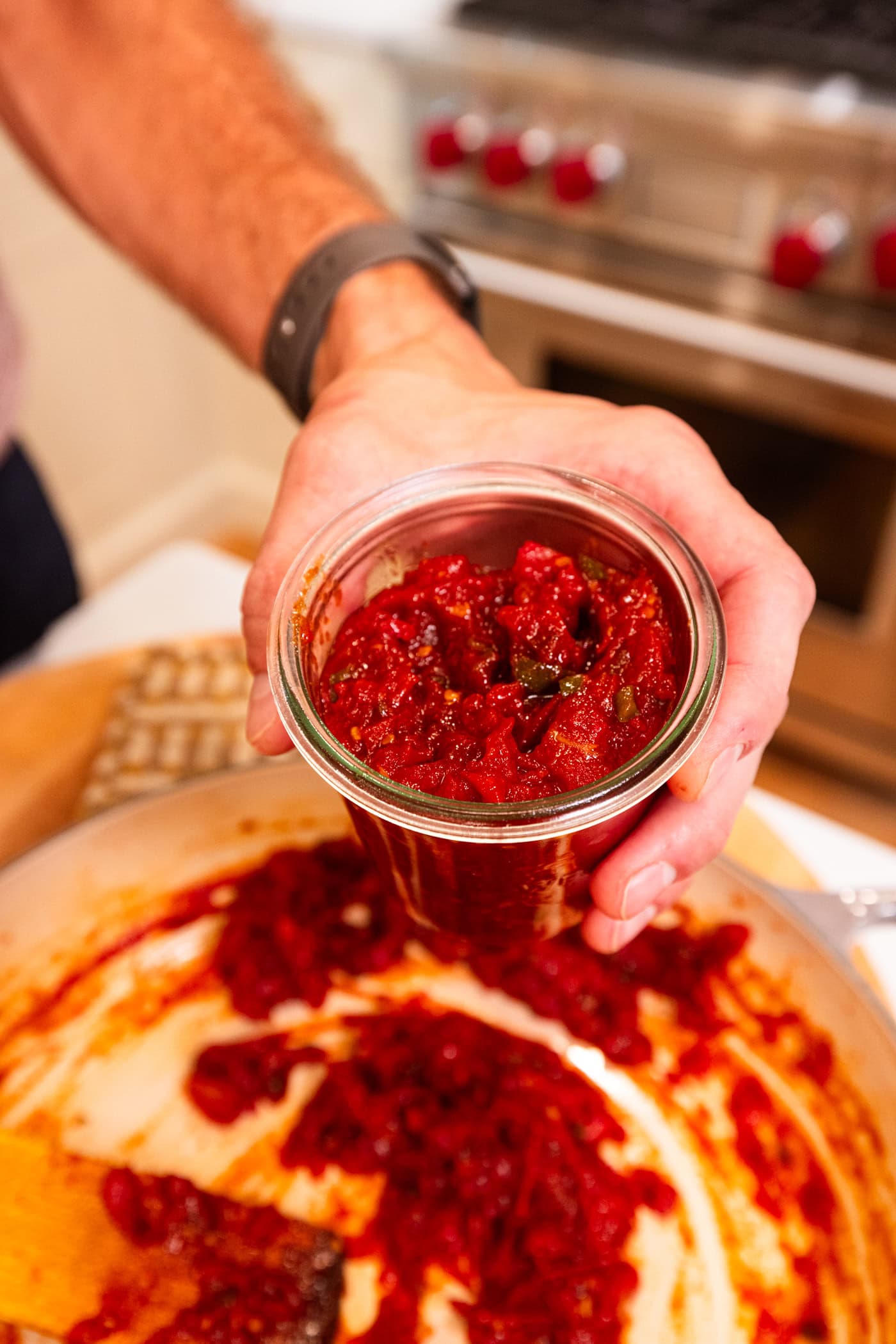 Hand holding jar filled with tomato jam after scooping it out of a skillet.