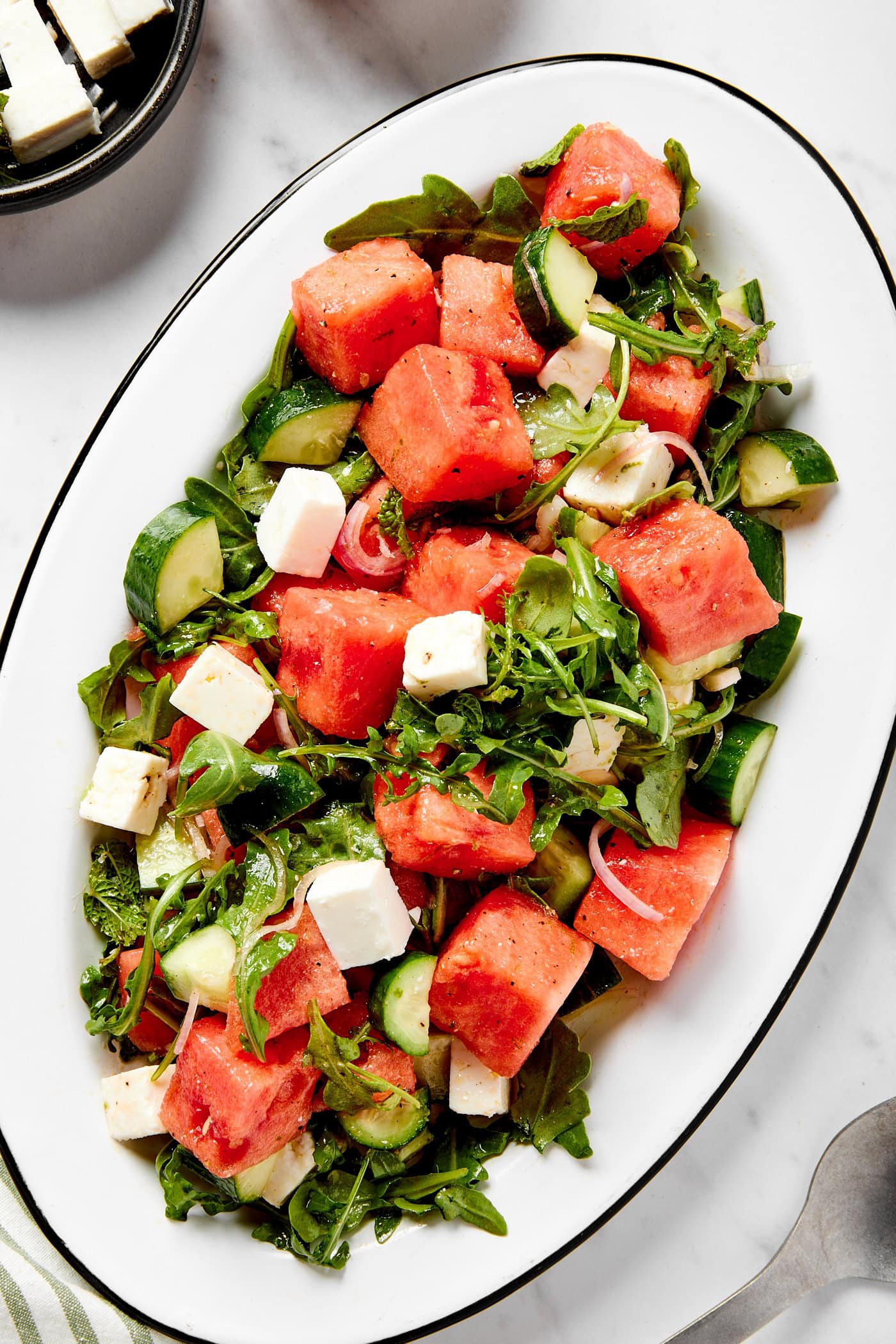 White serving platter with watermelon plated on top with cubes of feta and cucumber and arugula all on white marble surface.
