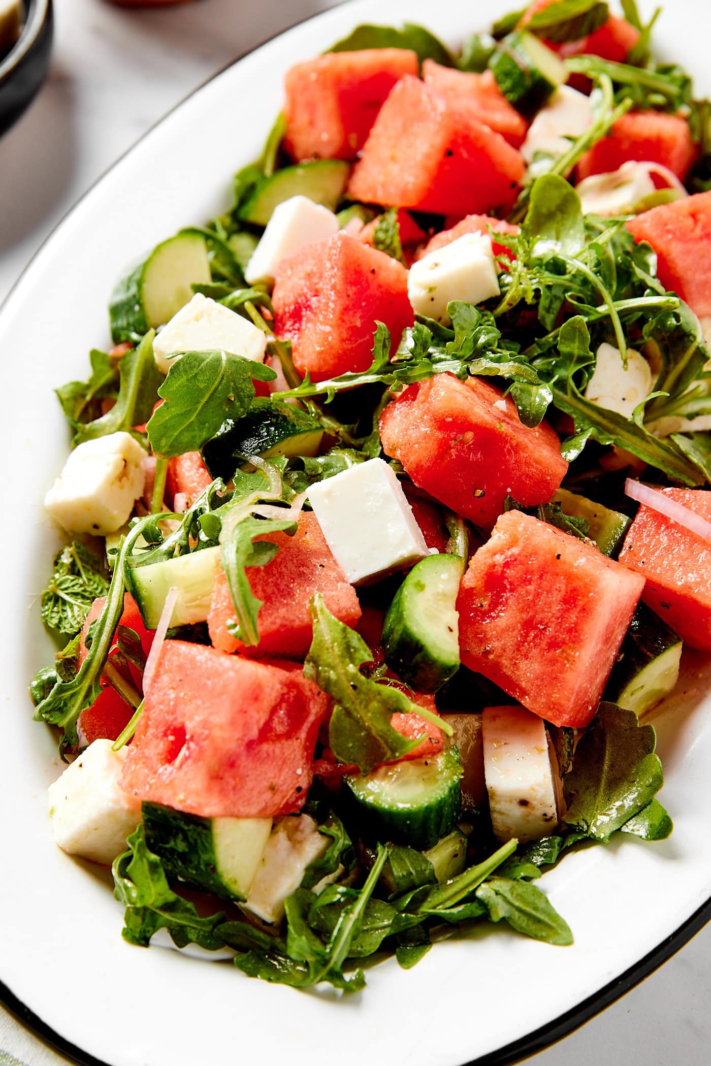 Chunks of bright pink watermelon sitting on a bed of arugula tossed with dressing with feta and cucumber on top as well.