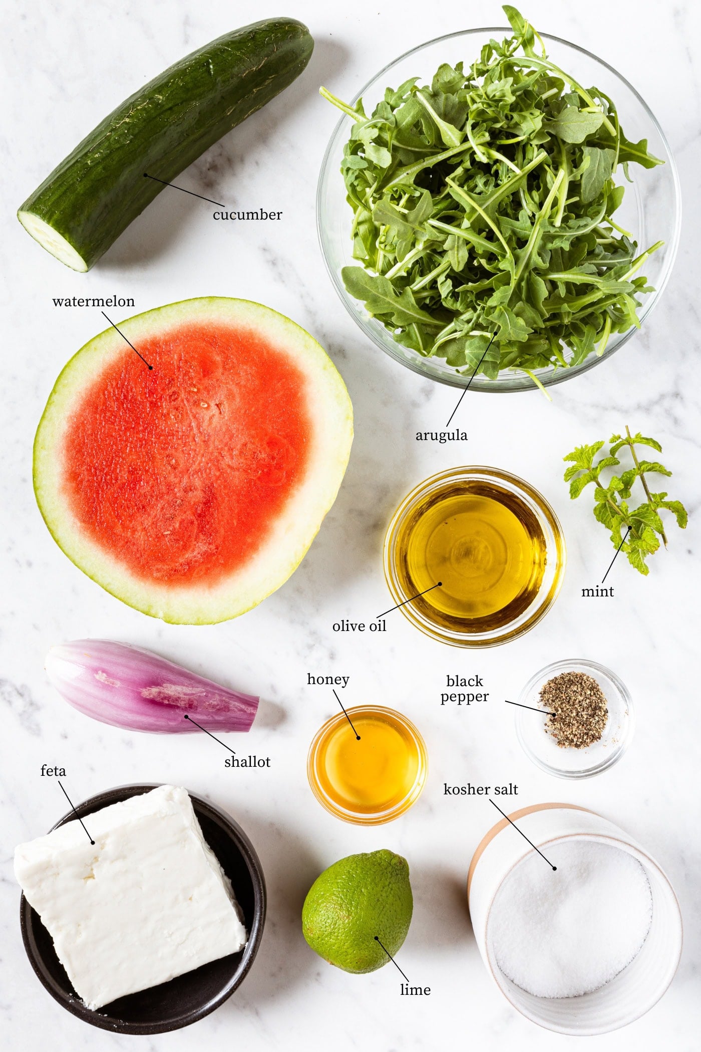 White marble surface filled with ingredients needed for salad including arugula, cucumber, watermelon, spices, and more.