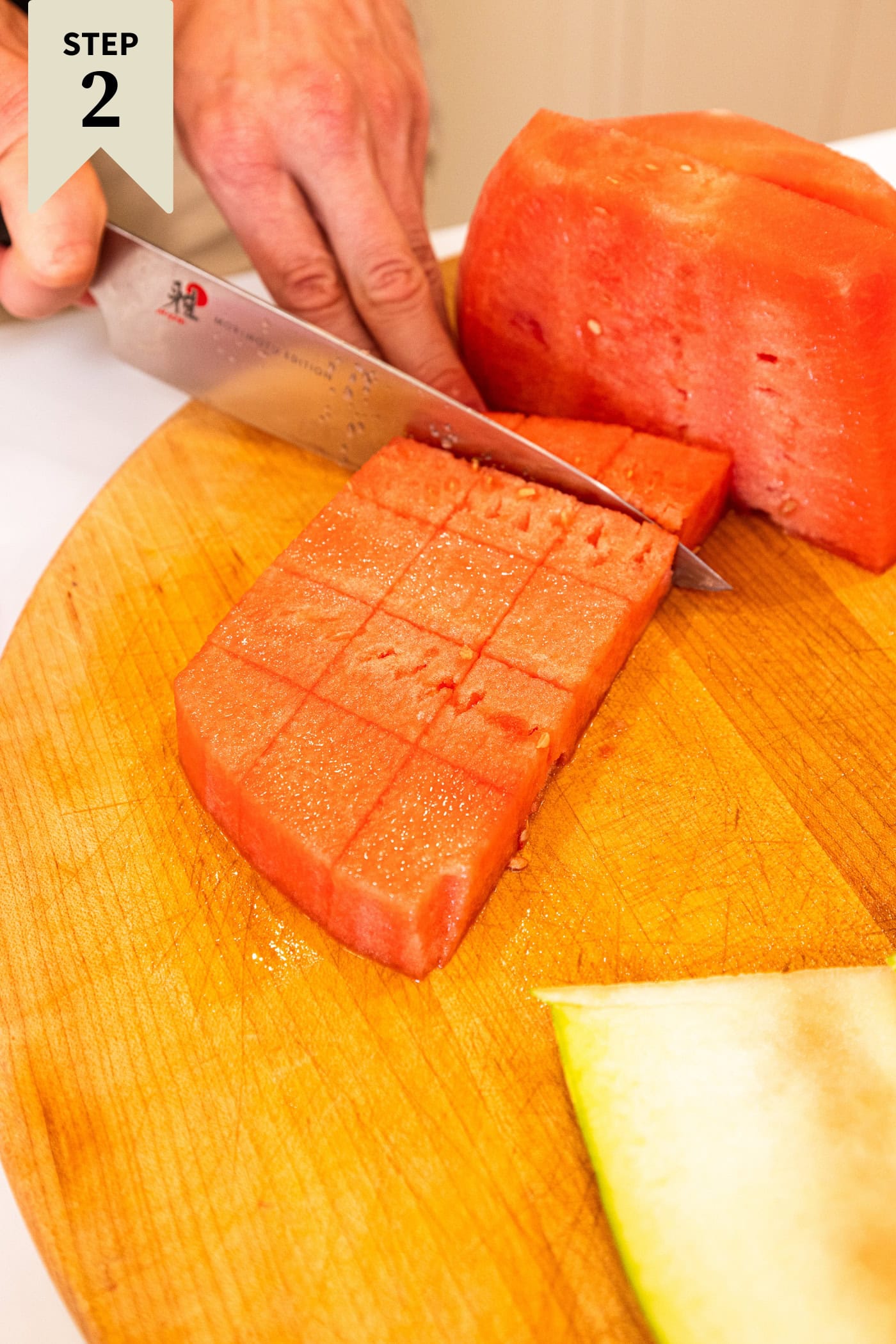 Hand using knife to slice watermelon into square cubes on a wood cutting board.