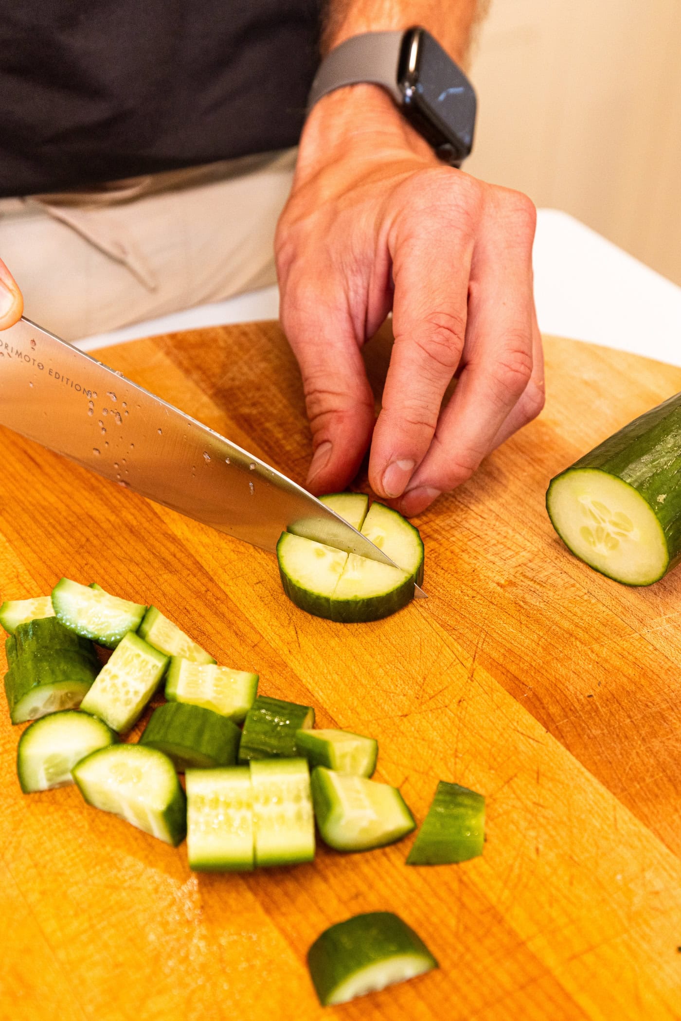 Hand using knife to slice cucumber into quarters on a wooden cutting board.
