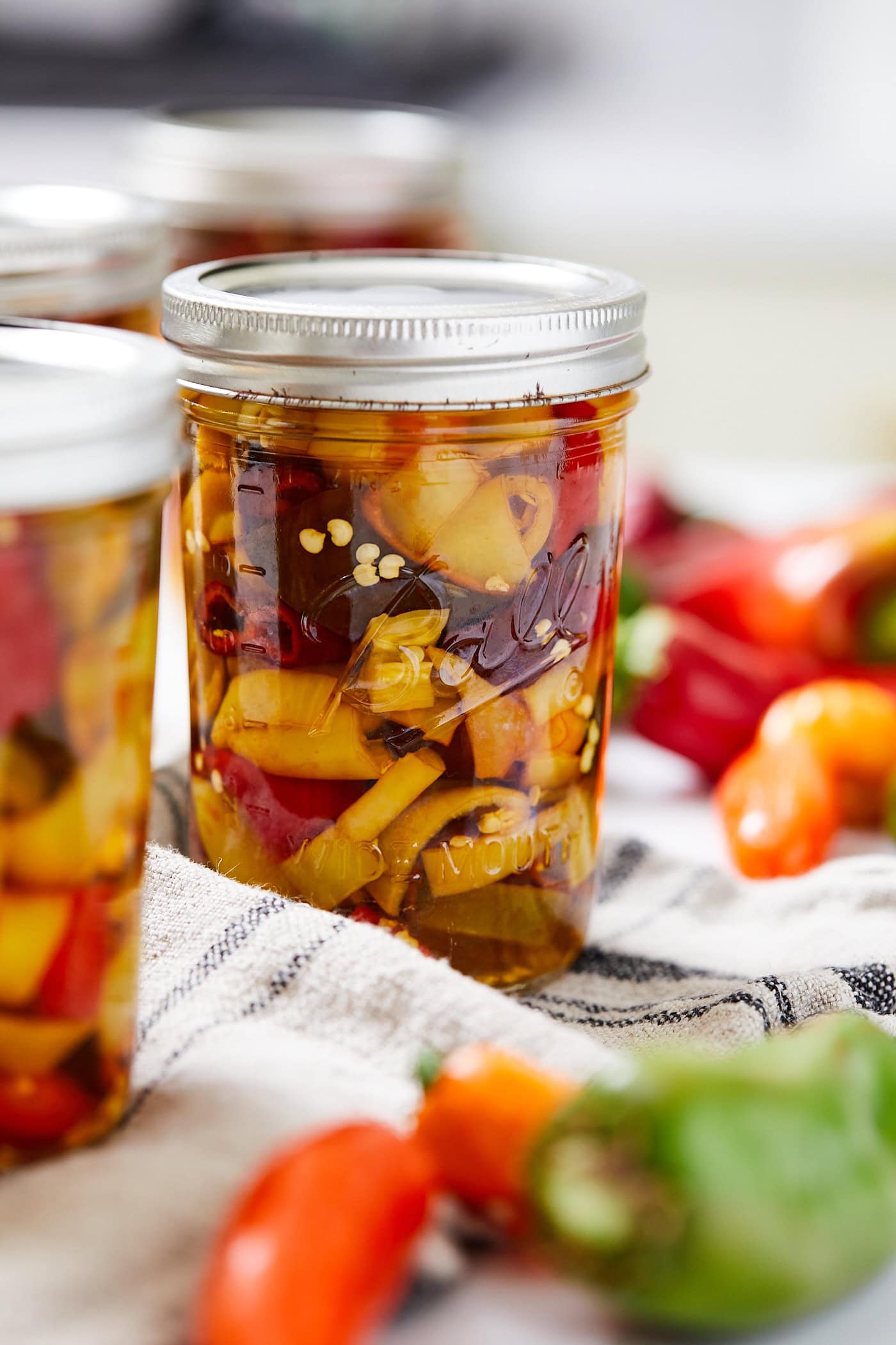 Glass jar with lid and ring on top with pickled peppers inside with seeds showing with more fresh peppers all around.