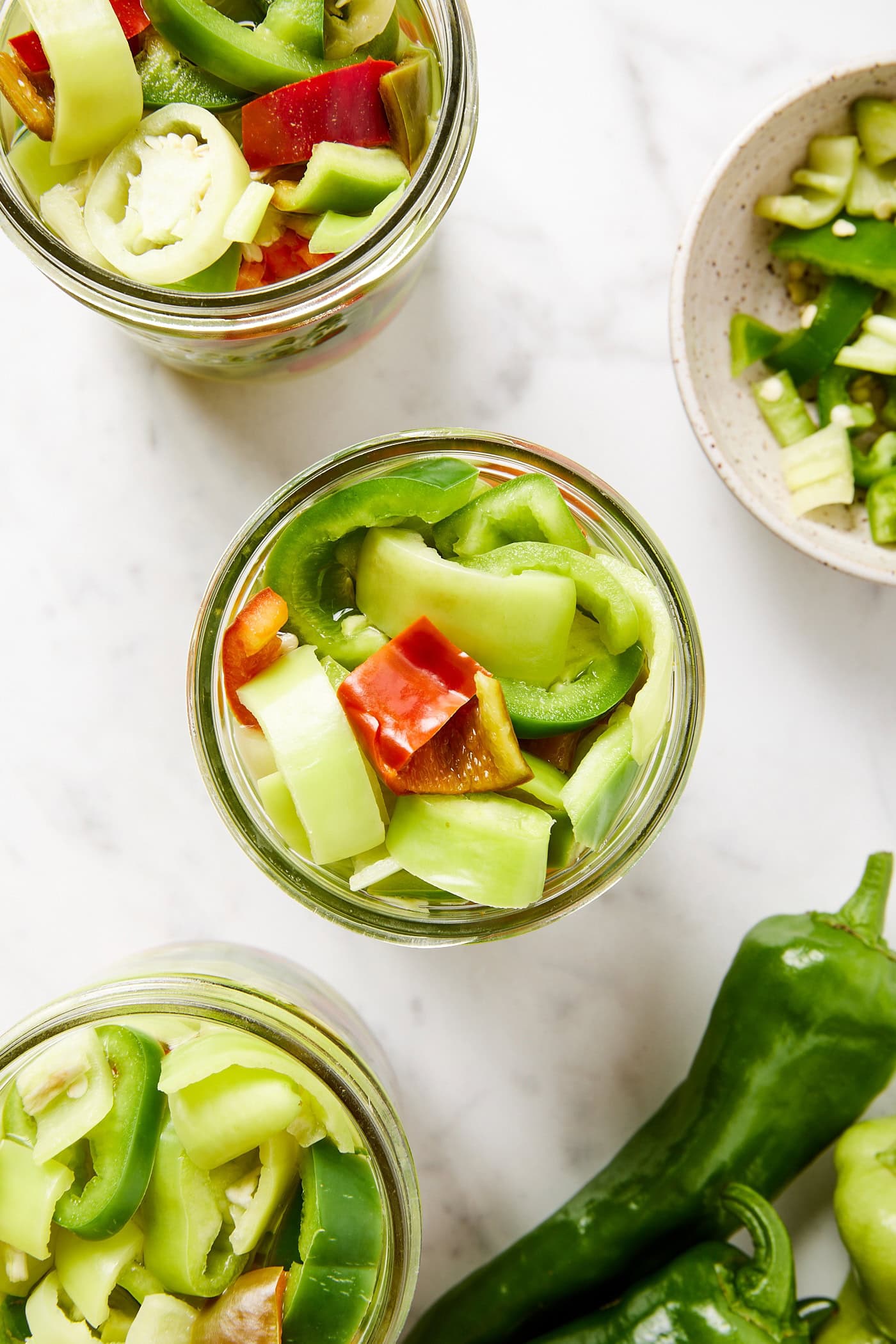 Glass jars filled with sliced peppers sitting on marble surface before brine is poured on top.