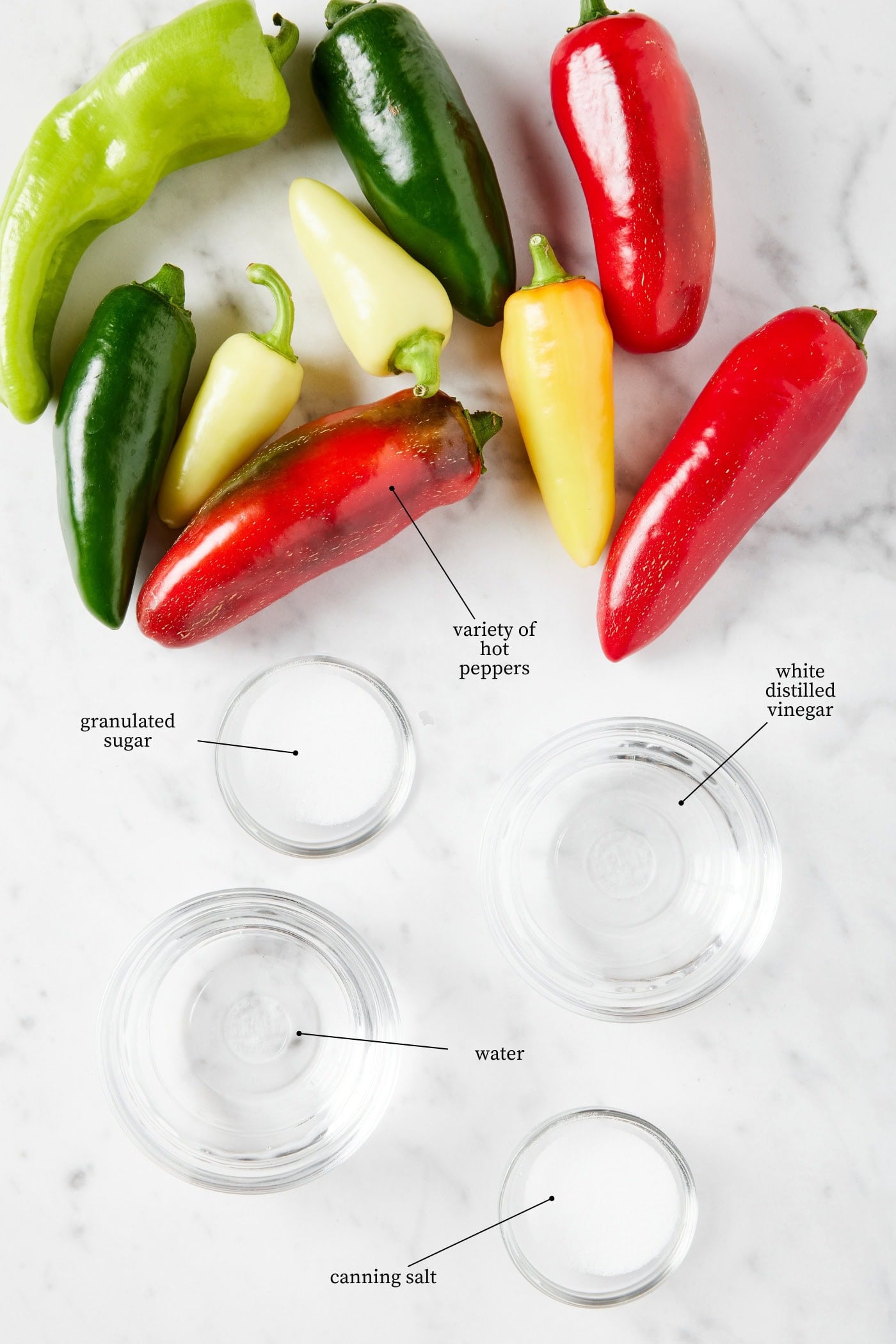 White marble surface with ingredients needed for canned pickled peppers including hot peppers, sugar, water, vinegar, and salt.
