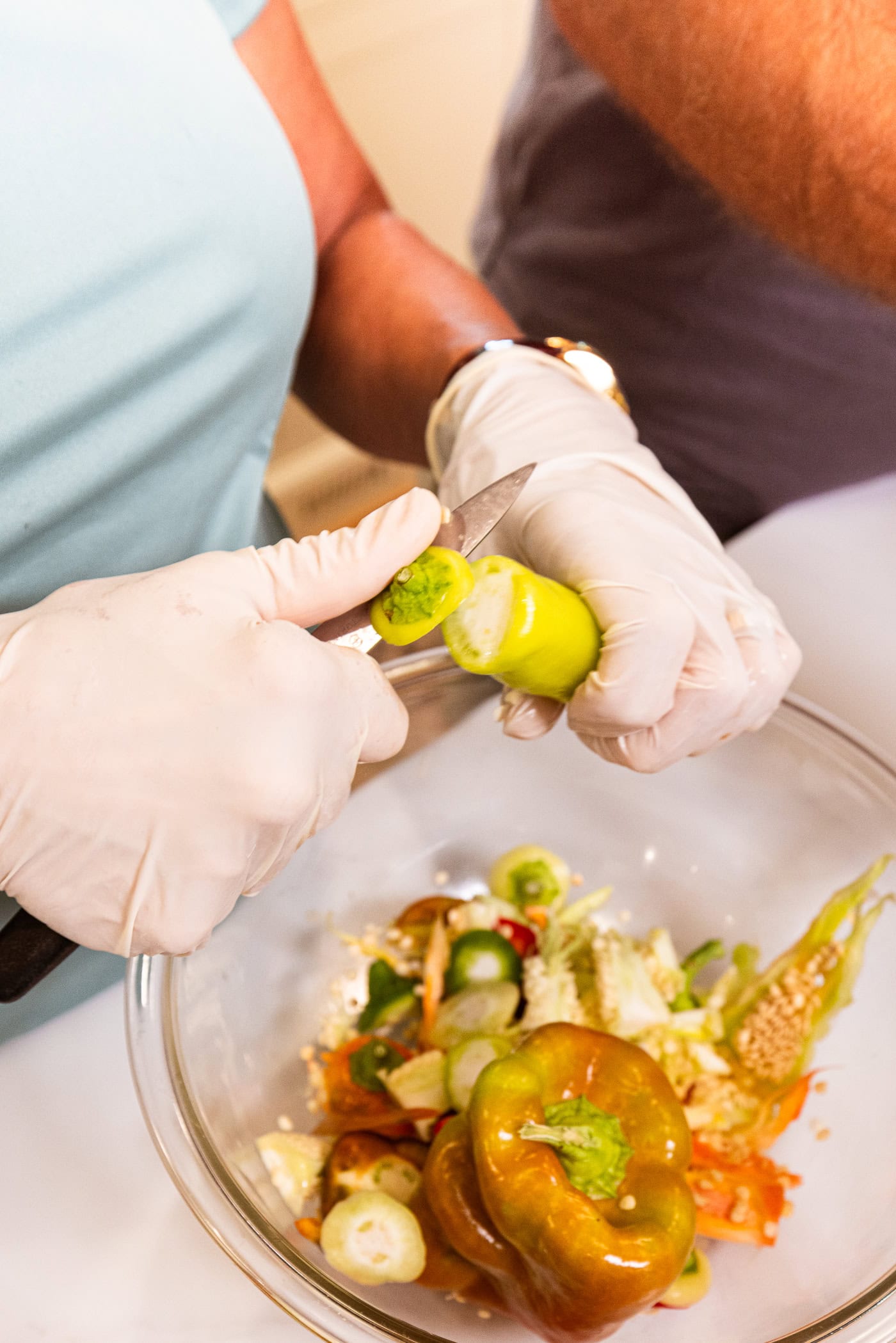 Two hands slicing off the end of a yellow hot pepper with discards in a bowl below.