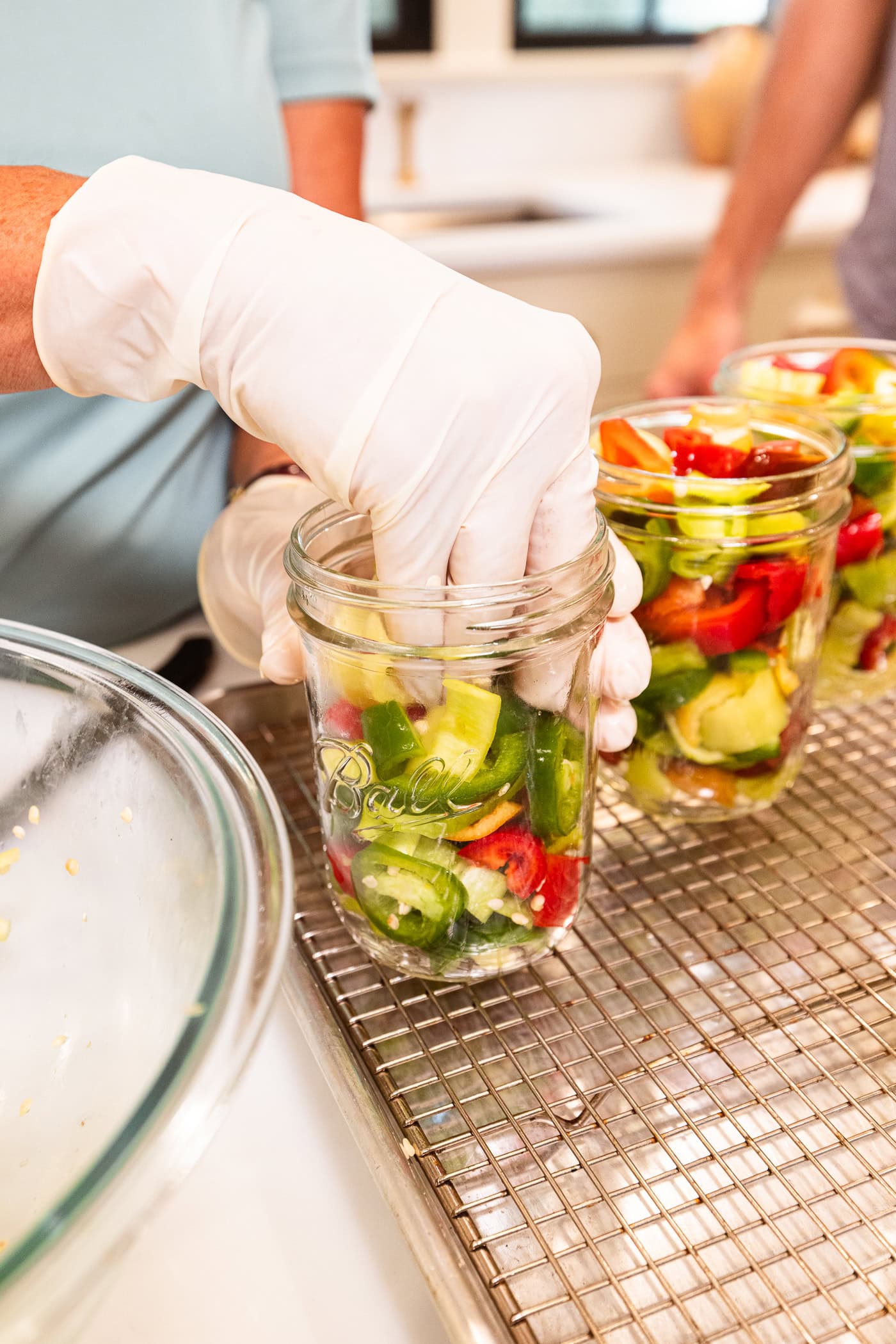 Hands wearing gloves packing slices of pickled peppers into a glass canning jar.