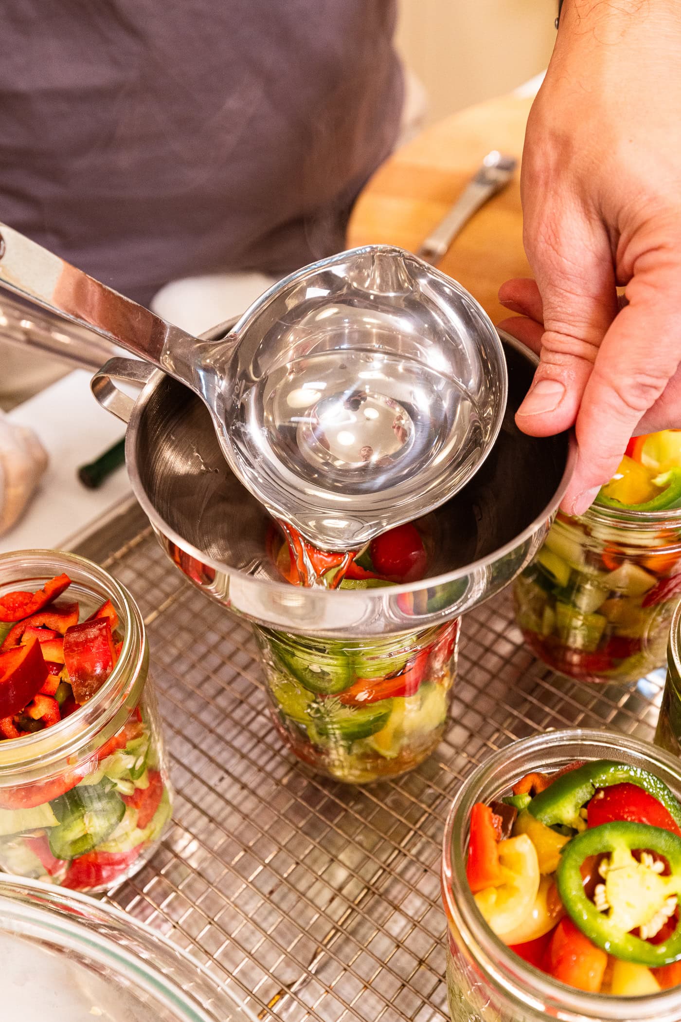Metal ladle pouring hot clear brine into a funnel set on a jar filled with sliced hot peppers.