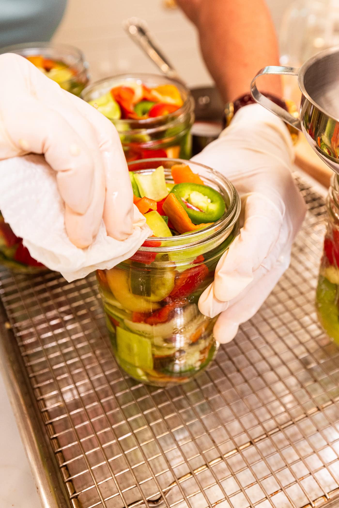 Gloved hand wiping off the rim of a glass canning jar filled with sliced peppers.