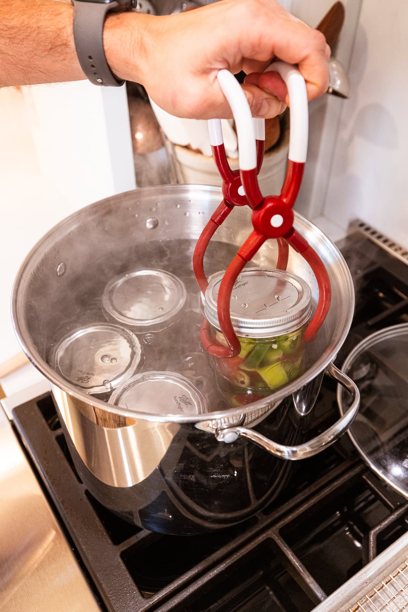 Hand using a jar lifter to place canning jars into a large metal pot filled with boiling water.