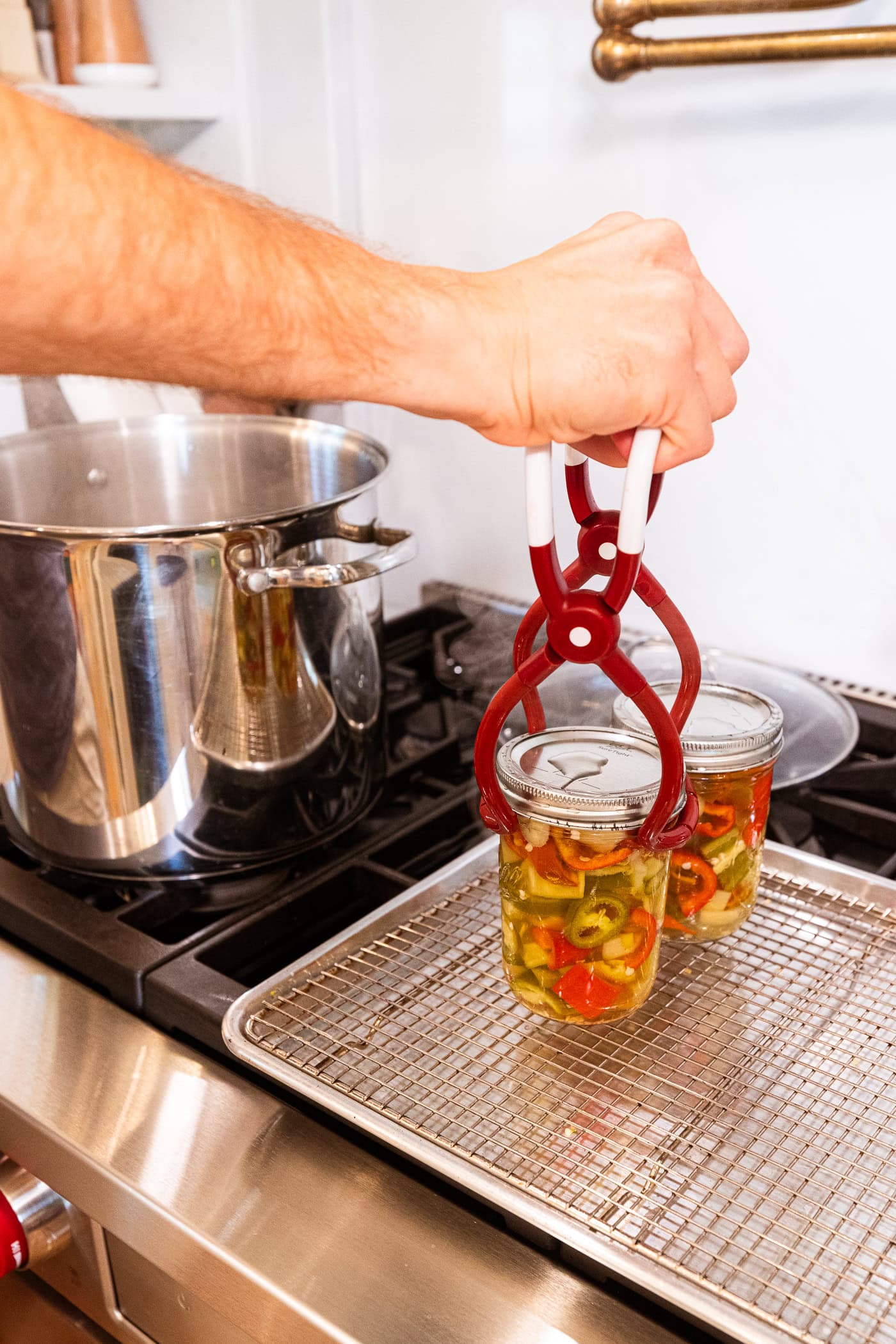 Hand using jar lifter to remove water bathed jars of pickled peppers onto a metal rack.