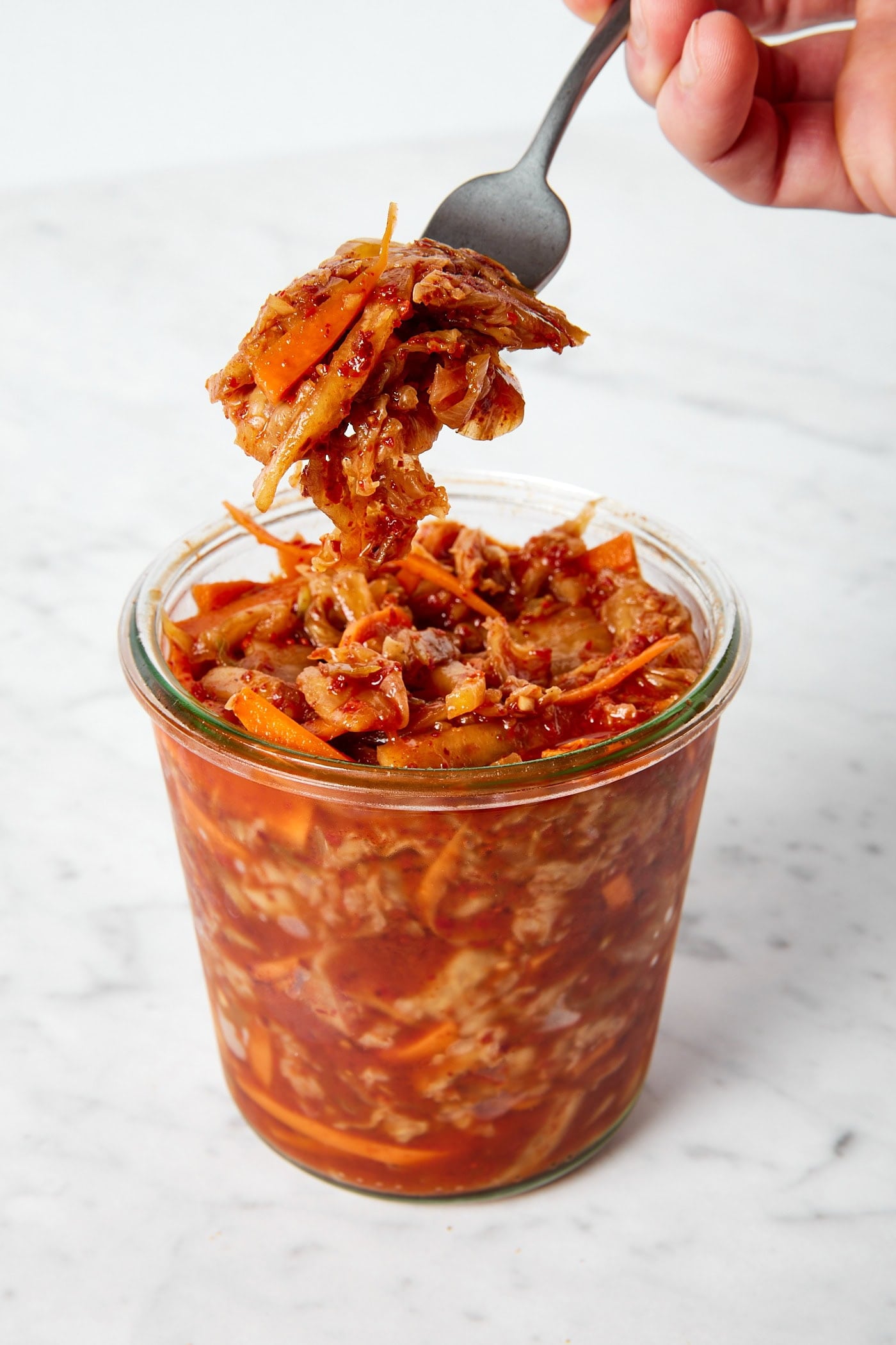 Fork picking up a serving of kimchi from a glass jar on a marble surface.