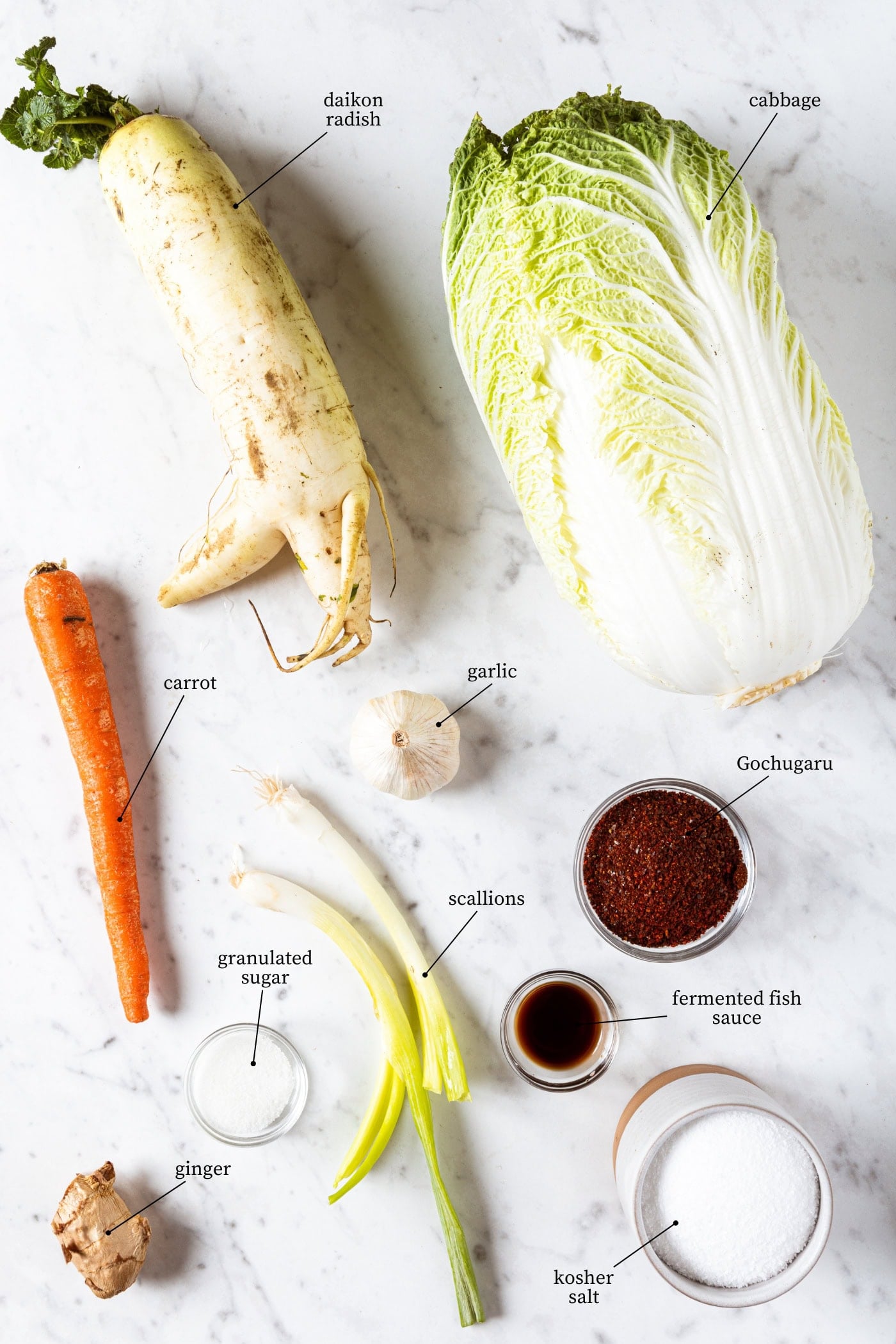 White marble surface filled with ingredients needed to make kimchi at home including cabbage, radish, fish sauce, salt, and more.