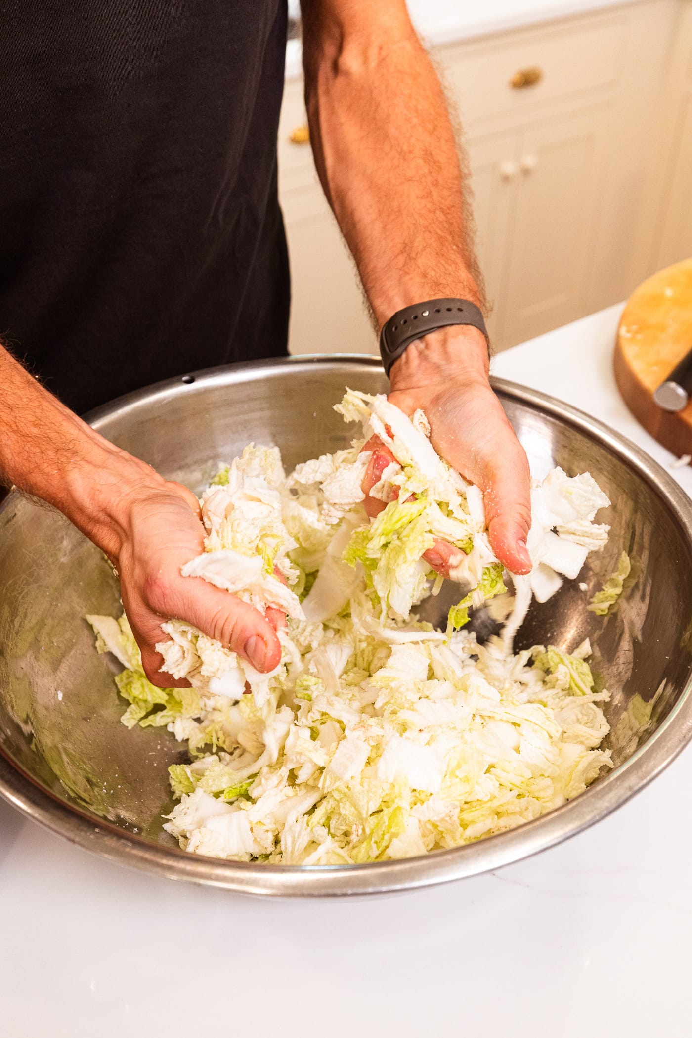 Two hands massaging sliced cabbage in a large metal bowl.