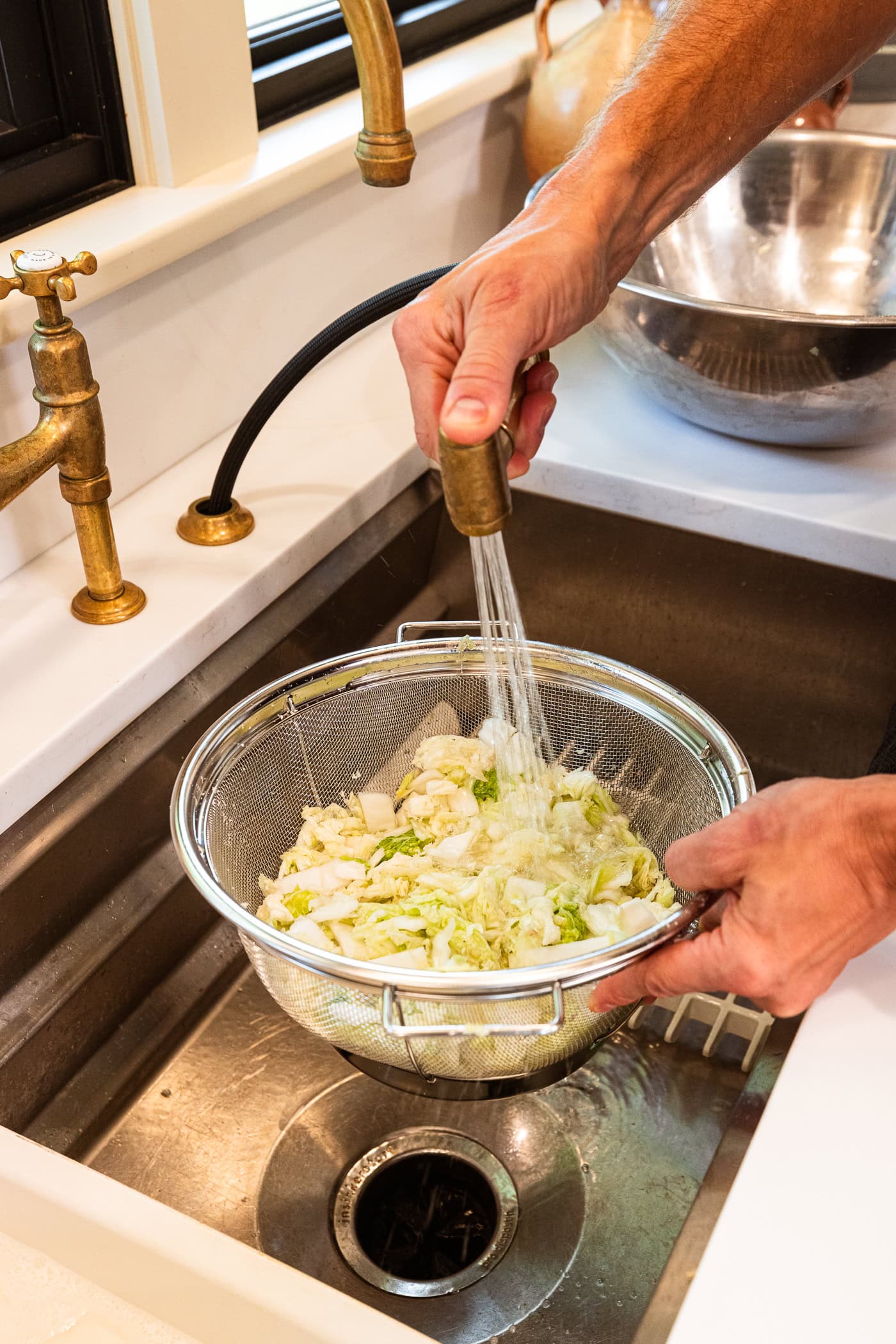 Hand using water faucet to rinse strainer filled with sliced cabbage in a sink.