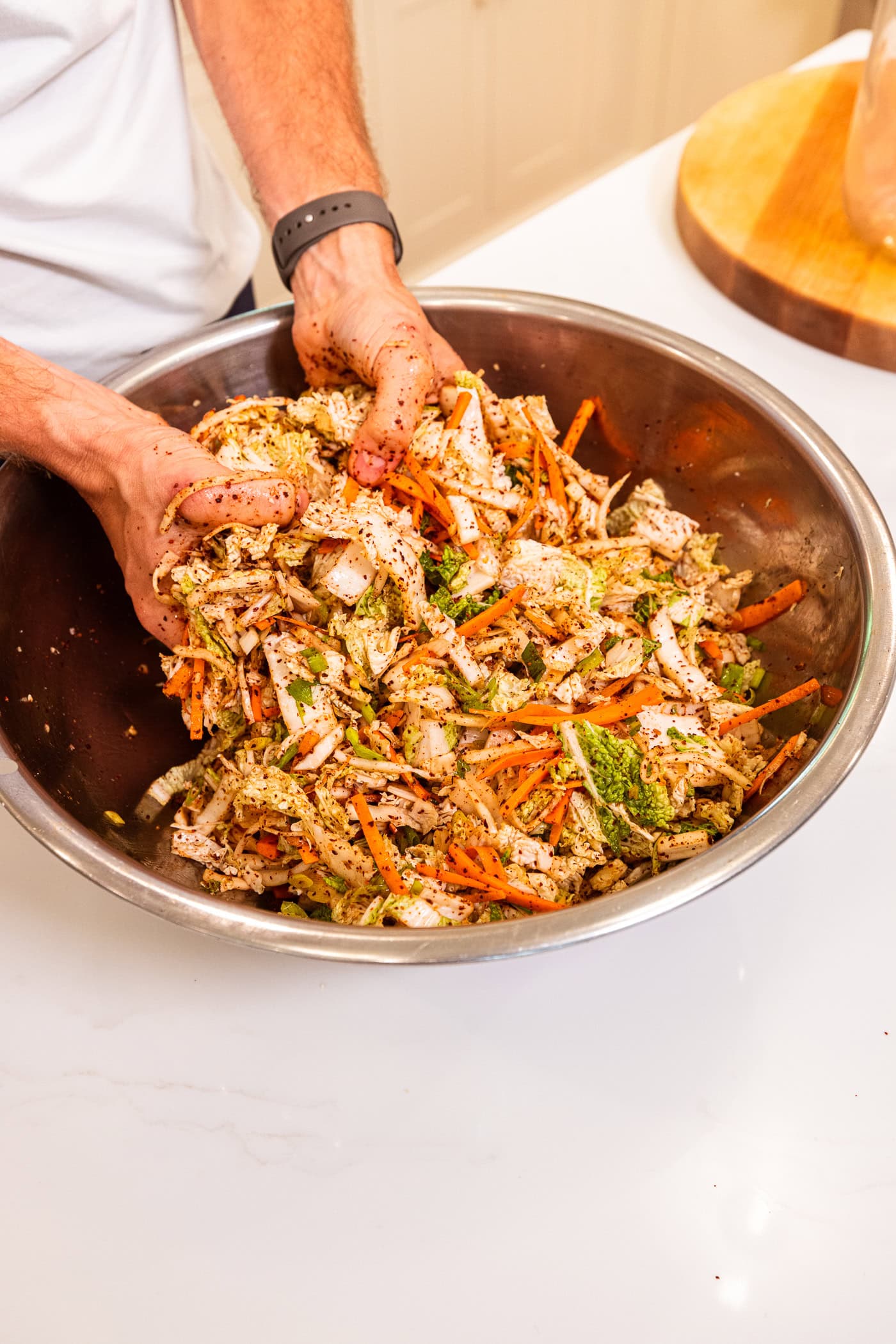 Hand mixing together spices with cabbage and other sliced vegetables in a large metal bowl.
