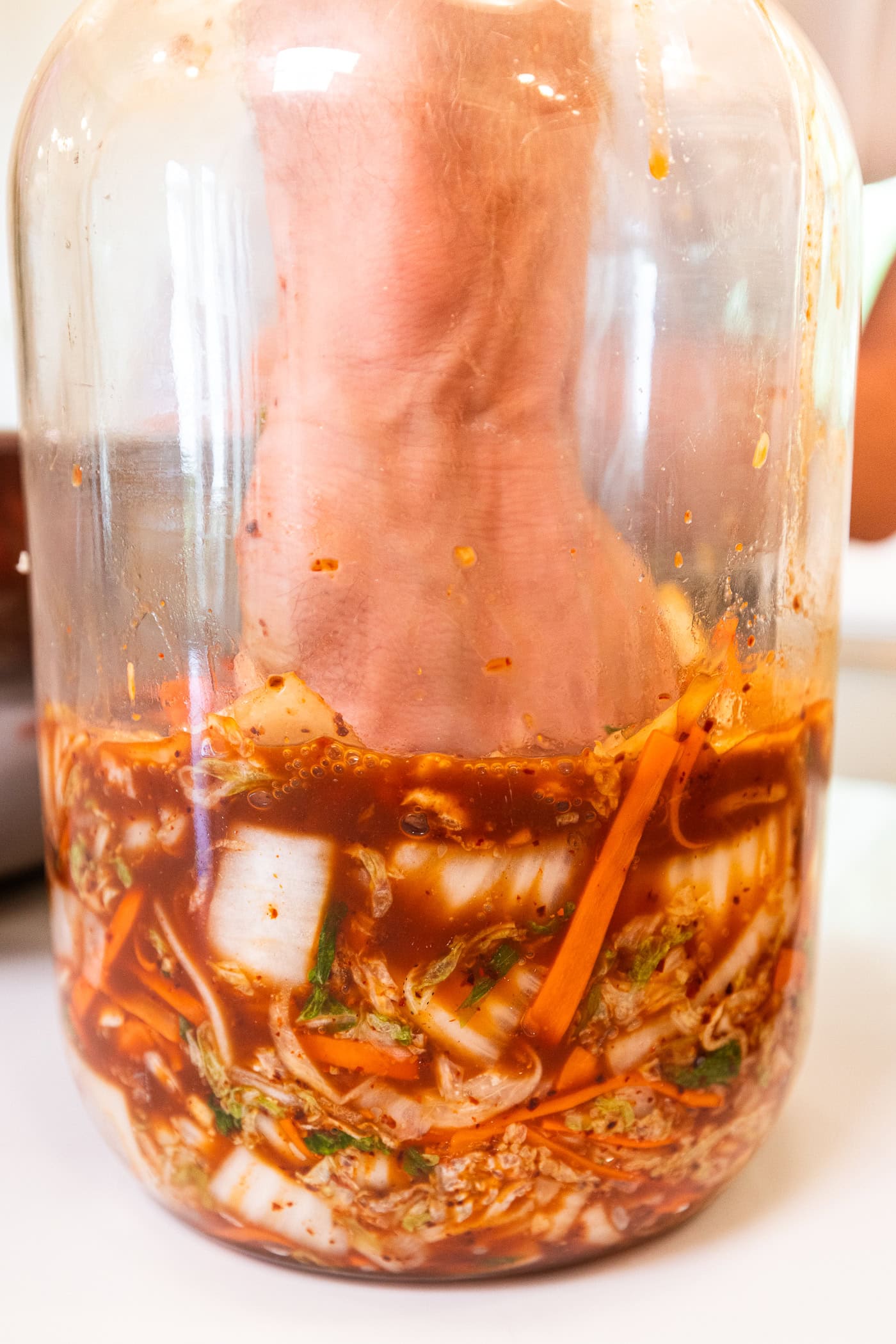 Hand pressing kimchi down into its orange liquid in a glass jar.