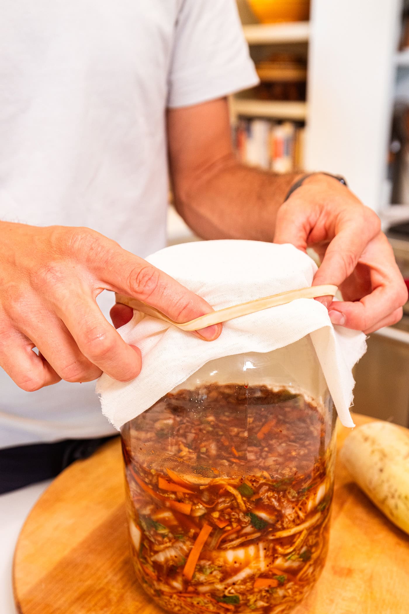 Hands placing rubber band on top of a glass jar with cheesecloth over the top.