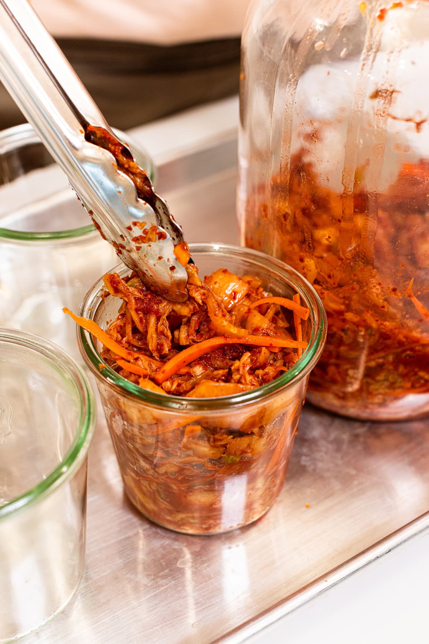 Tongs placing orange colored homemade kimchi into glass jars on metal tray.