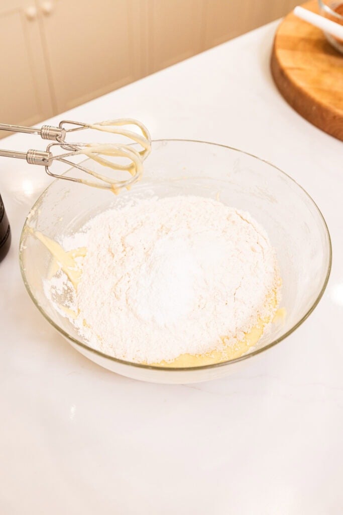 Flour sits on top of a wet batter, just about to be whisked by an electric hand mixer.