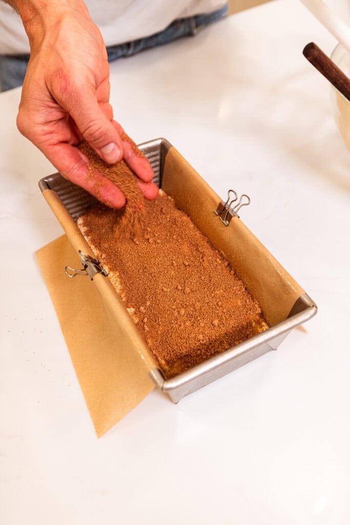 A hand sprinkles brown sugar crumble overtop coffee cake batter in a loaf pan.