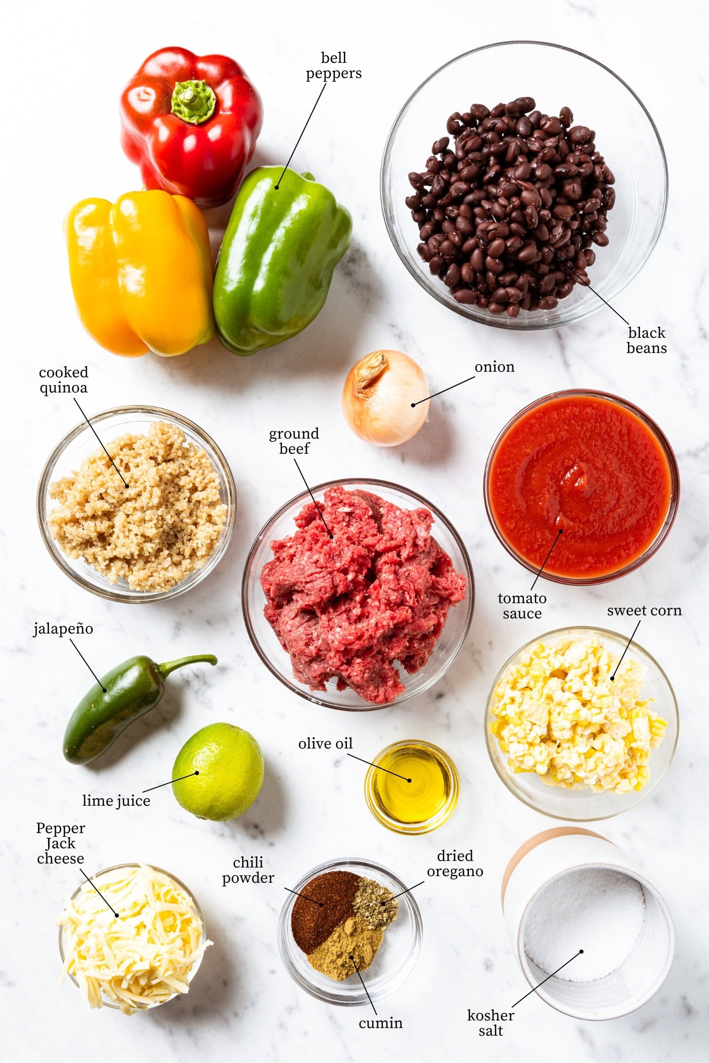 Ingredients needed to make stuffed peppers sit on a white marble surface including bell peppers, black beans, an onion, a jalapeno, quinoa, ground beef, tomato sauce, a lime, olive oil, corn, shredded cheese, chili powder, dried oregano, cumin, and kosher salt.