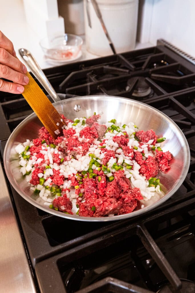 Ground beef, peppers, and onions cook in a skillet on a stove.