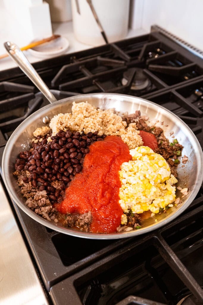 Tomato sauce, corn, black beans, and quinoa all sit on browned beef ready to be mixed in.