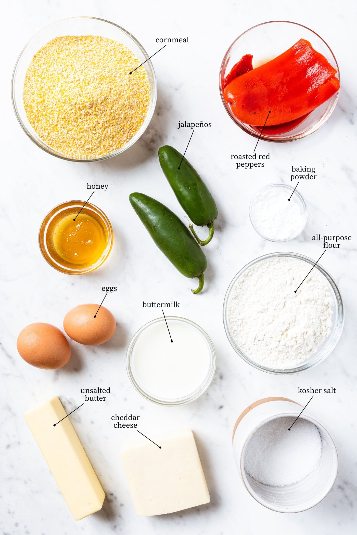 Ingredients for brown butter jalapeño cornbread sit on a marble surface. Ingredients include: cornmeal, red roasted peppers, jalapenos, honey, baking powder, flour, eggs, buttermilk, butter, cheddar cheese, and kosher salt.