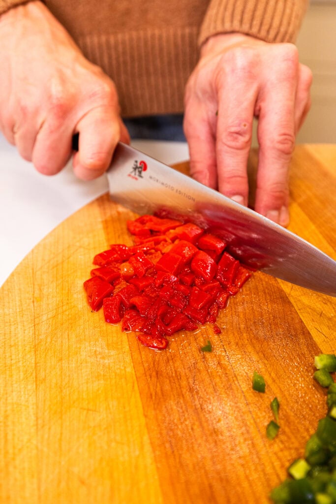 A hand dicing fire-roasted peppers on a wooden cutting board.
